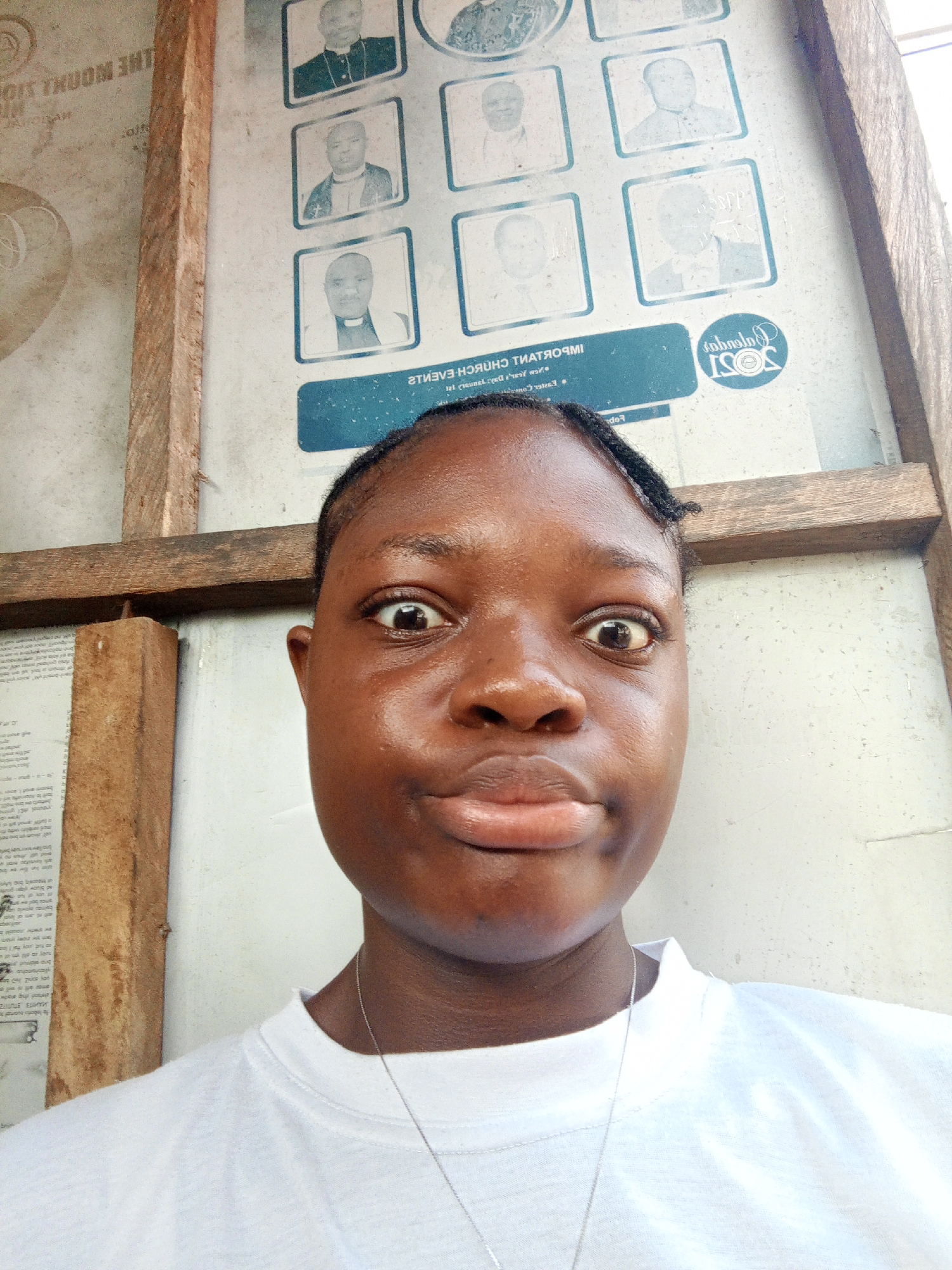 A close-up selfie shows a young Black person with dark skin, prominent eyes, and pursed lips, looking directly at the camera with a surprised or playful expression. Small braids frame their forehead. They are wearing a white crew-neck t-shirt and a thin silver necklace. Their face appears somewhat shiny.
The background features a light-colored wall or board, partially obscured by rough wooden beams that form a vertical and horizontal cross-section. On the board, several framed portraits of men are visible, rendered in a light blue/grey tone. Some of these men wear clerical collars, suggesting a religious context. Below the portraits, there's inverted text, part of which reads "IMPORTANT CHURCH EVENTS". A blue circular logo with "2021" and possibly "Calendar" is also visible. Further text, some fragmented or inverted, includes "THE MOUNT OF...", "Barouqay", "SOSI", and "CHURCH-EVENTS". On the far left, another section of text is affixed to the wall.
The setting, likely in Mbribit, Nigeria, appears to be an indoor or covered outdoor area, possibly within or near a church or community building, given the nature of the visible text and portraits. The lighting is soft and diffused, indicating daytime. The overall impression is informal and spontaneous.