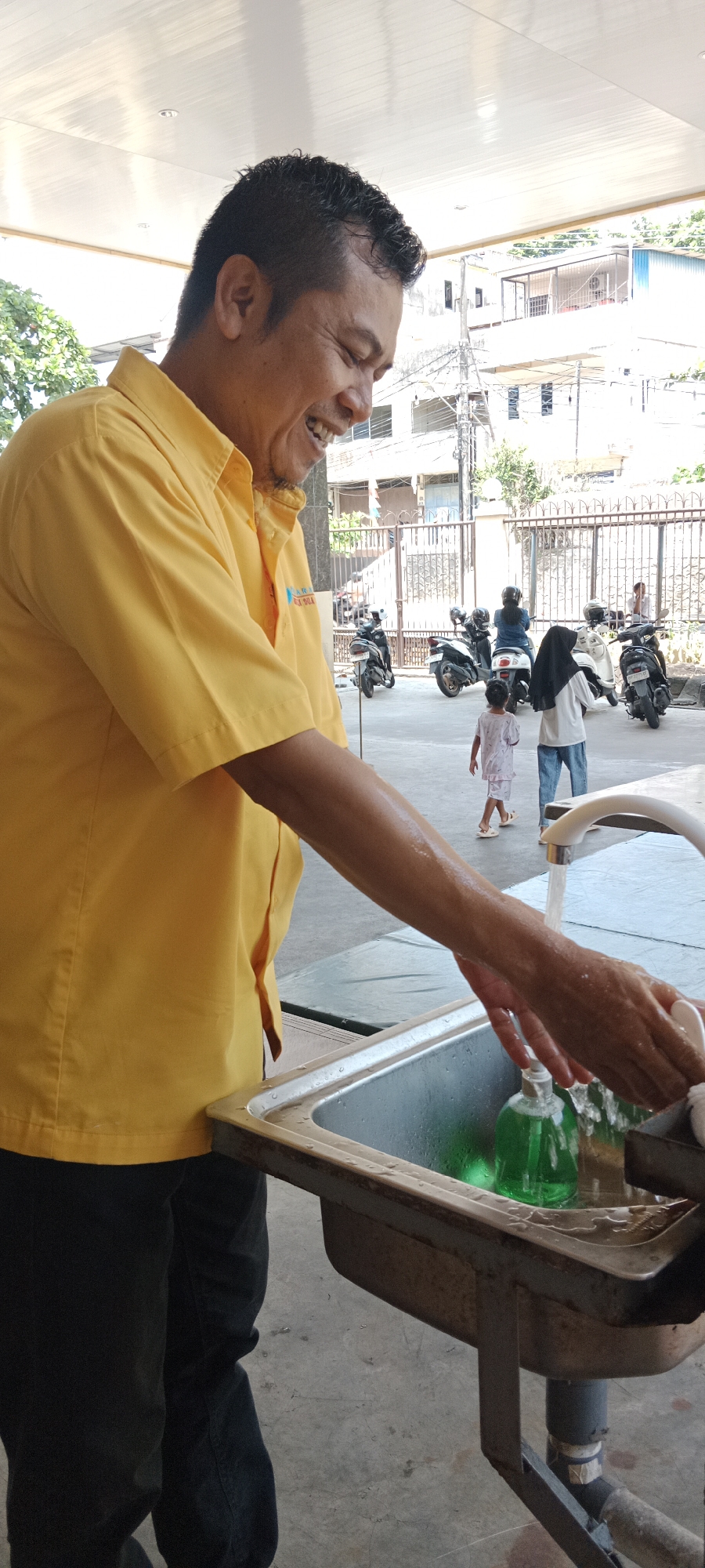 A man in a yellow collared shirt and dark pants is washing his hands at an outdoor sink in front of a building in Tanjung Pinang, Indonesia. He is smiling. The sink is made of metal and has a faucet with running water. There is a green bottle of soap in the sink. In the background, there are several motorcycles parked and a few people walking. The buildings in the background appear to be residential. The overall lighting suggests it is daytime.