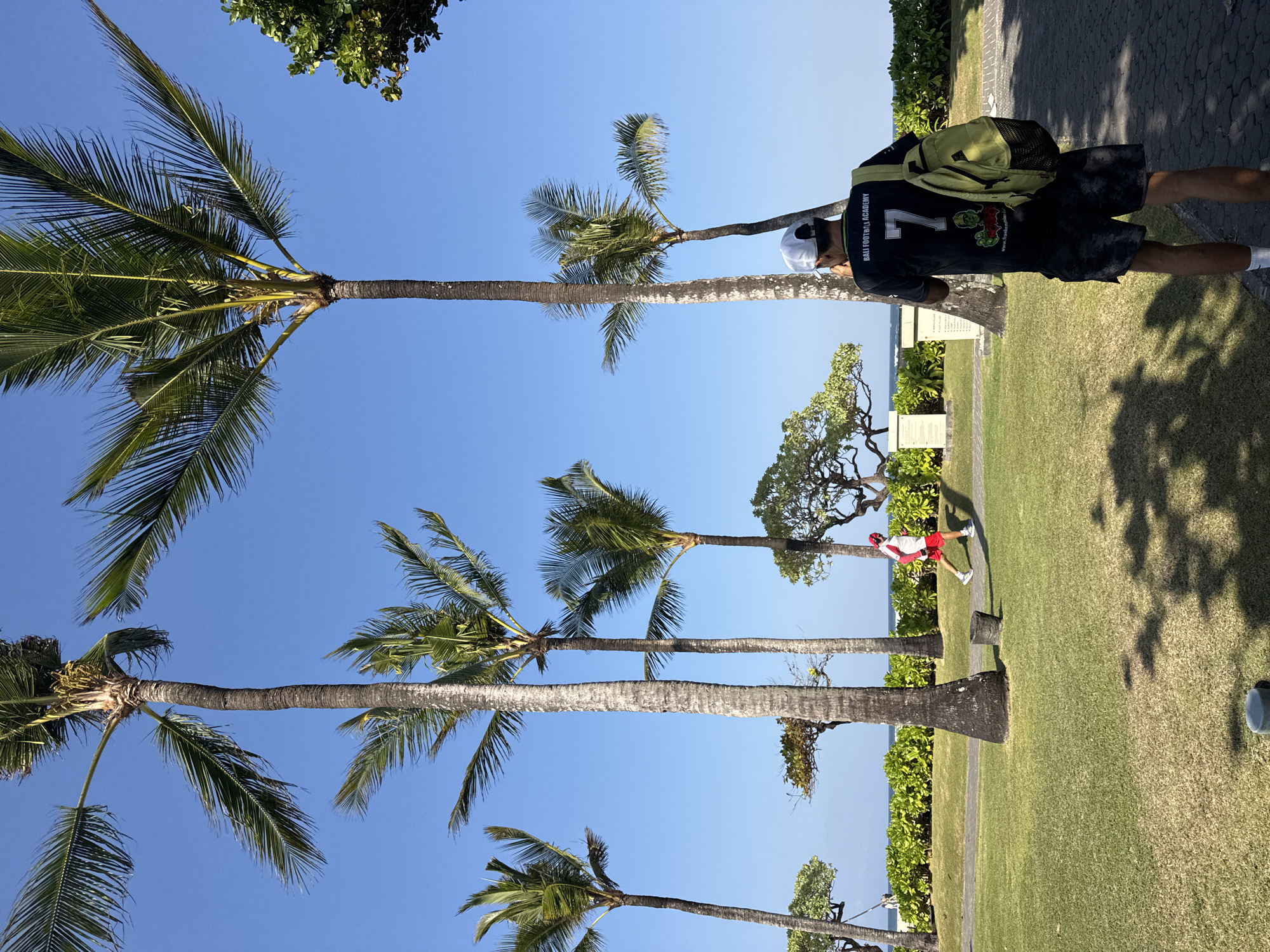 This image captures a scene in Kuta Selatan, Indonesia, on a bright, sunny day. The foreground features a man wearing a black t-shirt with the number 7 and "BALI FOOTBALL ACADEMY" printed on the back, along with dark patterned shorts, a white cap, and a light green backpack. He is walking away from the camera on a paved pathway. In the mid-ground, a person in athletic wear, including a red cap, white shirt, and red shorts, is jogging on a grass path. The background is dominated by tall palm trees with lush green fronds against a clear blue sky. A line of dense green bushes and vegetation runs along the edge of the property, separating it from a beach and the ocean in the distance. The overall environment suggests a tropical resort or park setting.