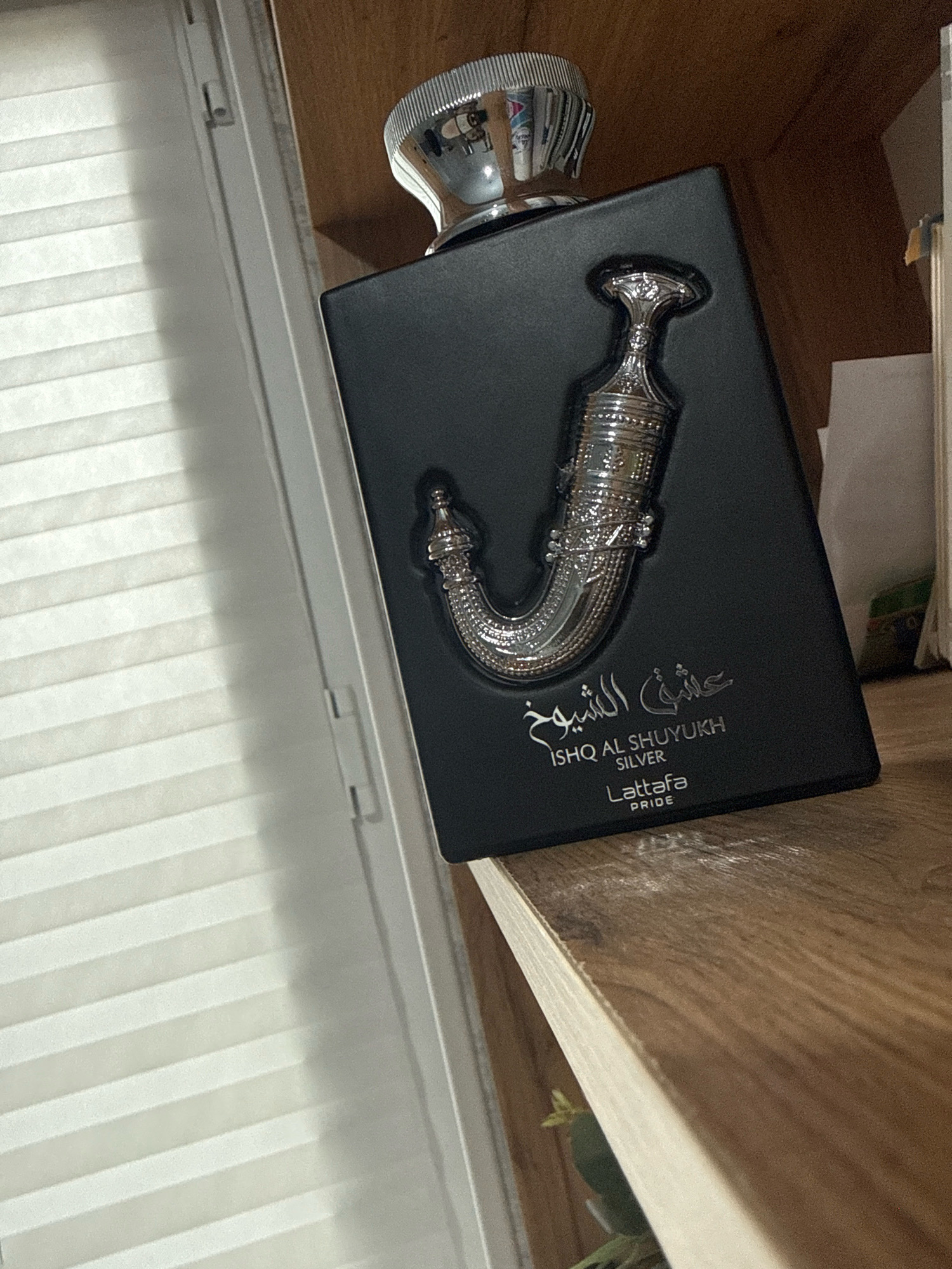 This is a close-up, eye-level shot of a black perfume bottle with silver embellishments. The bottle has a unique, ornate silver design resembling a khanjar dagger on its front. Arabic script and English text are visible on the bottle, reading "ISHQ AL SHUYUKH SILVER" and "LATTAFA PRIDE." The bottle is positioned on a wooden shelf, which appears to be part of a bookshelf. To the left of the bottle, a white window blind with horizontal slats is visible, casting shadows on the bottle. The background consists of the wooden shelf and a portion of a white wall or partition. The scene appears to be indoors. The city of Nazran, Russia is not depicted in the image.