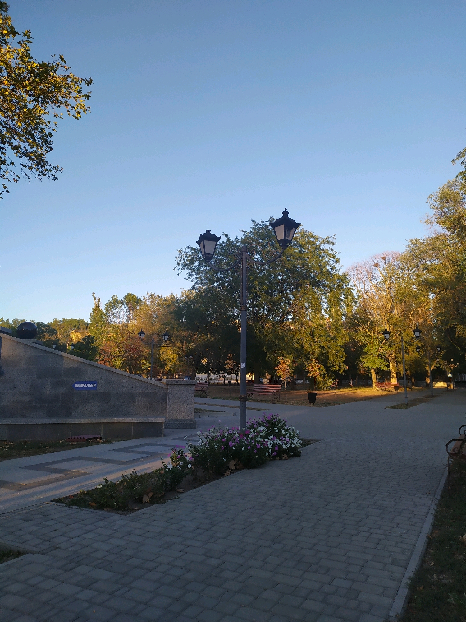 The image depicts an outdoor scene in Mykolaiv, Ukraine, likely a park or public square, during the daytime. The sky is clear and blue, suggesting fair weather. A paved walkway made of grey rectangular bricks runs diagonally across the lower right portion of the frame. To the left of the walkway, there is a flower bed with a mix of white and pink flowers.
Further to the left, a large, grey stone structure with a polished surface is visible. On this structure, a blue sign with white Cyrillic lettering reads "ВБИРАЛЬНЯ," which translates to "TOILET" or "RESTROOM." A black sphere is mounted on top of the stone structure. Beyond this structure, trees with green and yellow foliage suggest the presence of autumn.
Several lampposts with ornate black metalwork are scattered throughout the scene, illuminating the path. Benches are visible in the background, indicating a space for public relaxation. The overall impression is one of a well-maintained public space.
