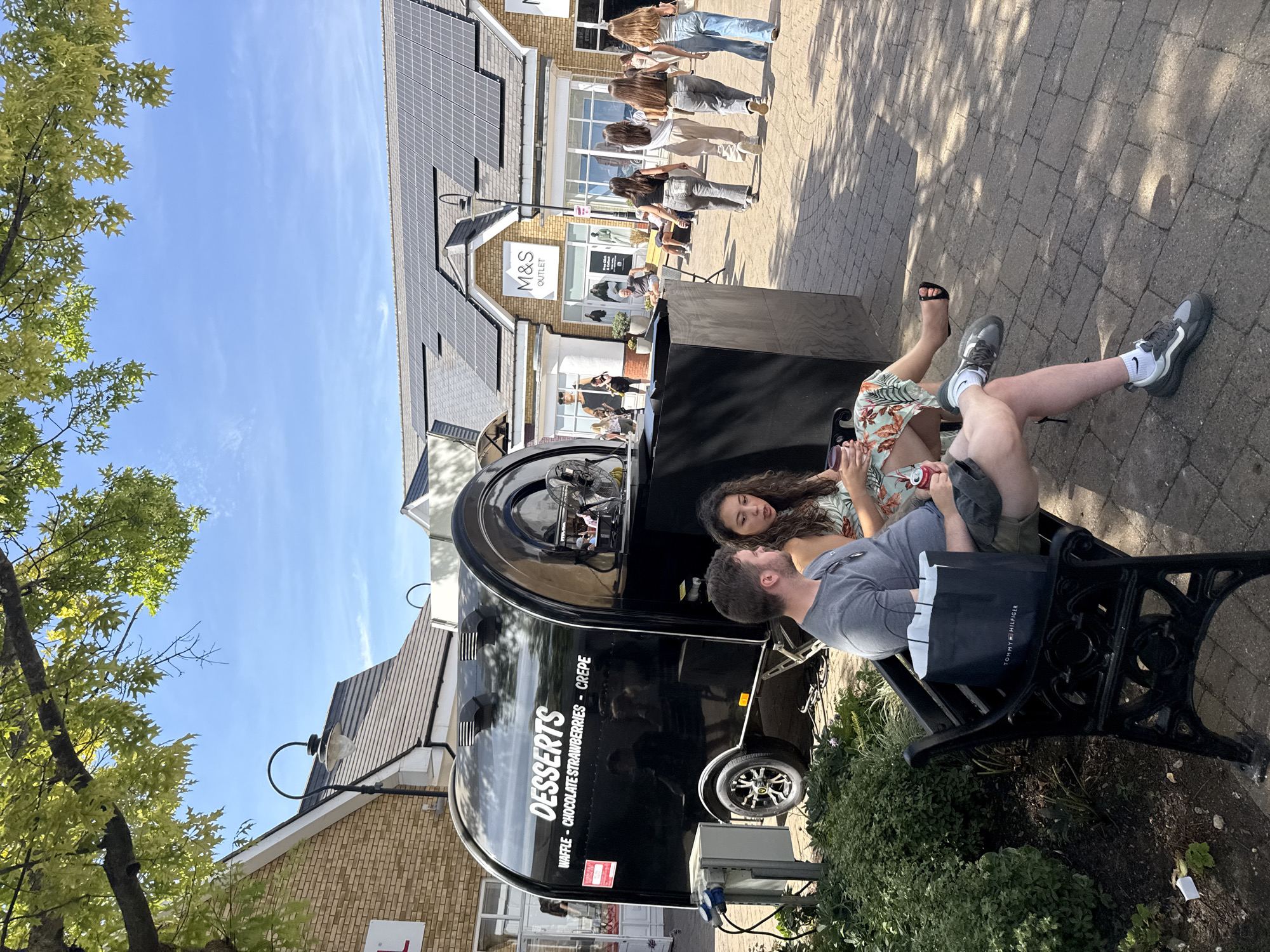 An exterior, medium shot captures a sunny day at a retail outlet in Braintree, United Kingdom. The image is centered on a black dessert trailer, advertising waffles, chocolate strawberries, and crepes. In the foreground, a couple sits on a black metal bench in front of the trailer. The man, wearing a grey t-shirt and shorts, looks towards the trailer, while the woman, in a floral dress, gazes at a cup in her hand. A Tommy Hilfiger shopping bag rests on the bench next to the man. Behind the trailer, other shoppers are visible walking in front of shops, including an "M&S Outlet" store. The ground is paved with light-colored bricks, and trees with green leaves provide shade. The sky is clear and blue with a few wispy clouds.