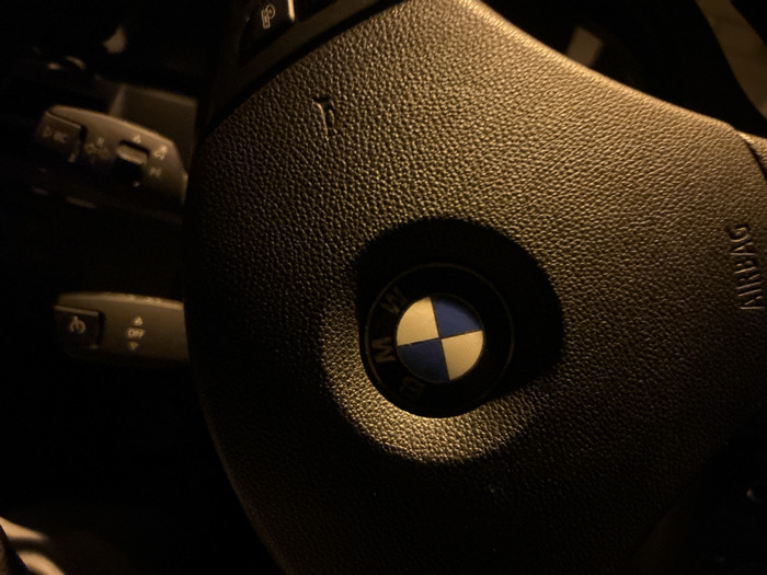 The image displays the interior of a vehicle, located in Voluyak, Bulgaria. The central subject is a dark, textured steering wheel, characteristic of a BMW, featuring the brand's blue and white circular emblem in its center. The word "AIRBAG" is embossed vertically on the right side of the steering wheel. To the left of the steering wheel, two control stalks are partially visible. The upper stalk displays symbols associated with lighting or turn signal controls, and the lower stalk shows "OFF" along with additional symbols, likely for wiper or cruise control functions. The scene is illuminated by dim, warm-toned light, creating high contrast and shadows, indicating a low-light environment. No individuals, actions, or interactions are present or depicted.