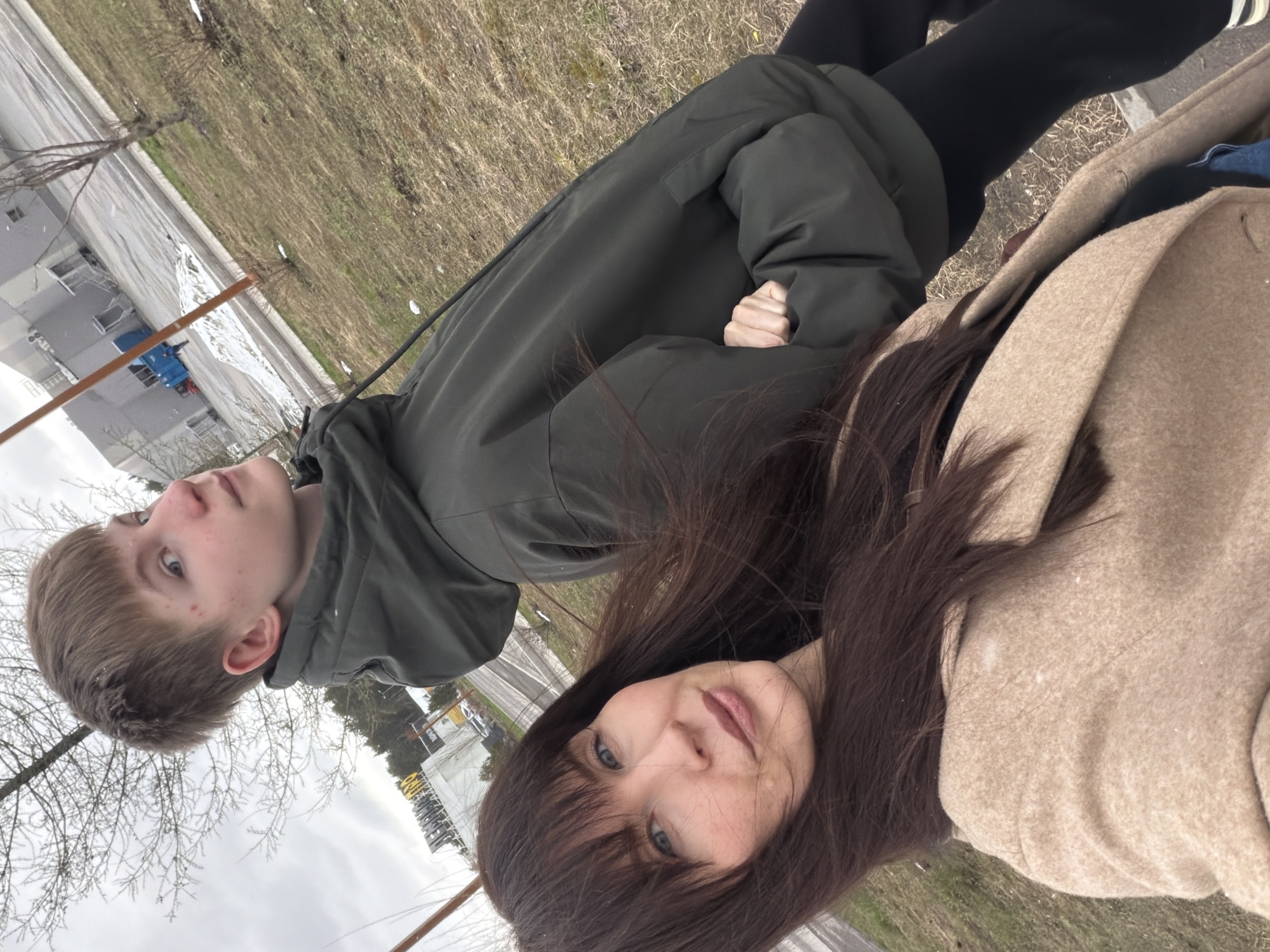 Two individuals, a young male and an adult female, are pictured outdoors. The male is wearing a dark green hooded jacket and black trousers, while the female is wearing a tan coat. The setting appears to be urban, evidenced by buildings and infrastructure in the background. Based on the location metadata (city of Perm, Russia), the image was likely taken at that location. The sky is overcast. The individuals are posing for the picture.
