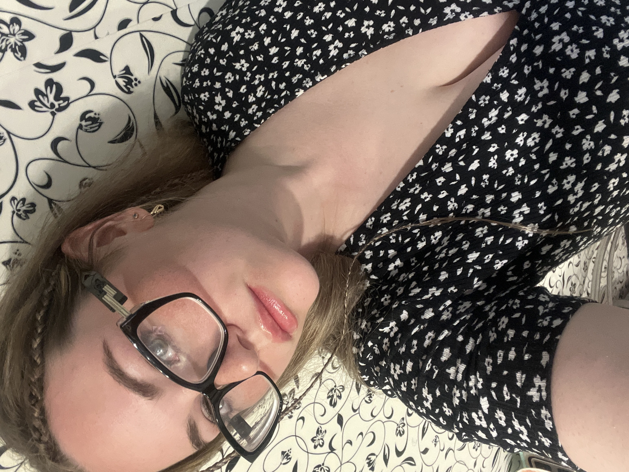 A young woman is lying down, angled towards the camera. She is wearing black-framed glasses and a black dress with a small white floral pattern. Her hair is light brown, with a braided section along her forehead. She has fair skin and is wearing lip gloss. She is lying on a white surface with a black floral pattern. It is not possible to confirm the location is Chaikovsky, Russia.