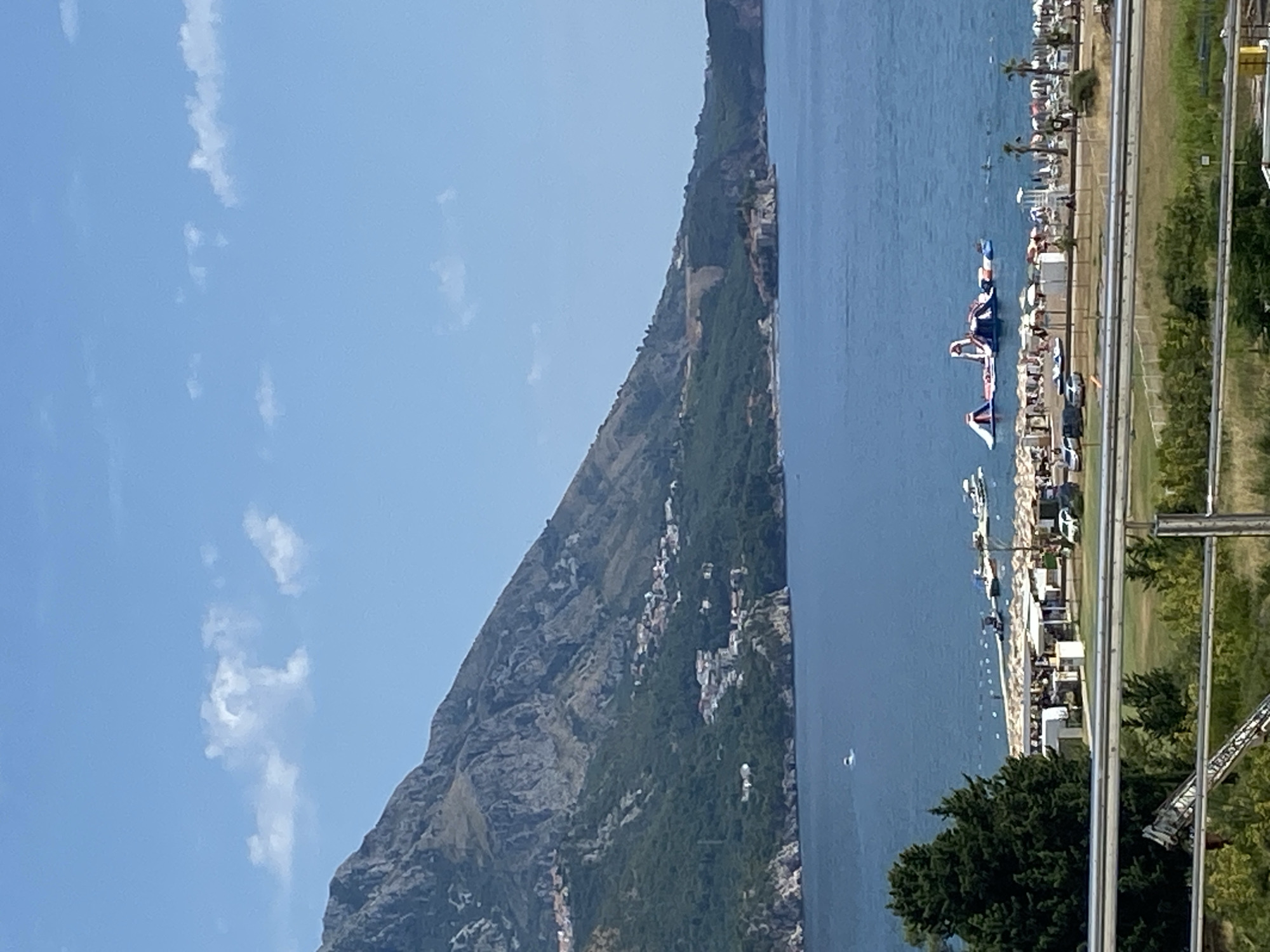 The image captures a scenic view of the Adriatic Sea coastline in Boreti, Montenegro. In the foreground, a metal railing and lush green vegetation frame the scene. Beyond this, a sun-drenched beach stretches along the coastline, dotted with numerous umbrellas and lounge chairs, indicating a popular tourist destination. Several white tents and structures are visible on the beach, likely serving as amenities for visitors. A few cars are parked near the beach.
Further out on the calm, blue sea, a large inflatable water park with slides and platforms is set up, with several people visible in and around it. A few small boats are anchored or moving near the shore.
The backdrop of the scene is dominated by a majestic, steep mountain covered in dense green forest. Small clusters of buildings, characteristic of Mediterranean architecture, are scattered along the lower slopes of the mountain, indicating a small settlement. The sky is a clear, bright blue with a few wispy white clouds. The overall impression is one of a vibrant coastal resort town on a beautiful summer day.
