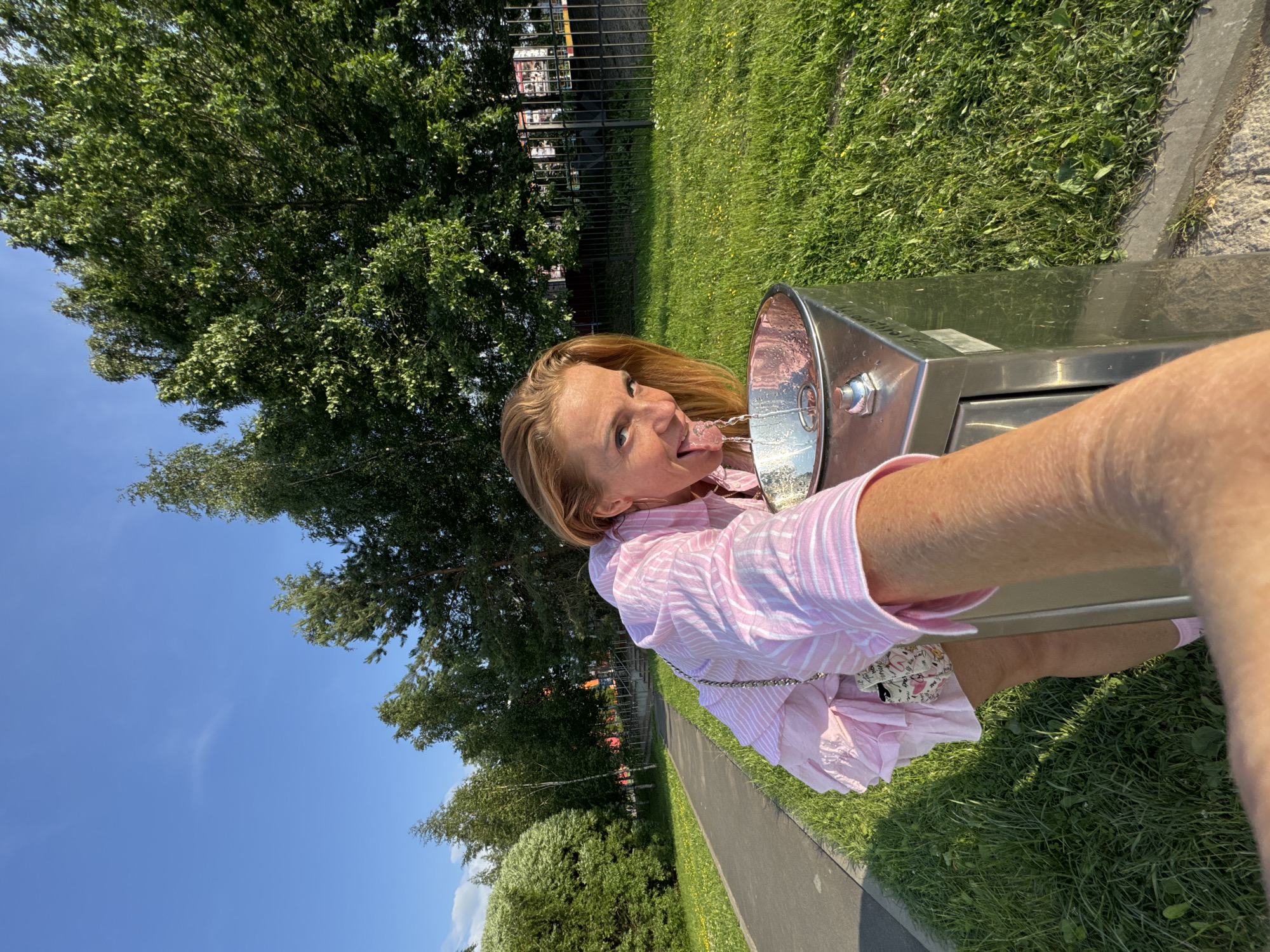 In Kirishi, Russia, a woman with blonde hair and fair skin, wearing a pink and white striped button-down shirt dress and pink socks, leans over a silver drinking fountain, sticking her tongue out to catch the stream of water. She is holding her left arm out, presumably holding a camera, as the image appears to be a selfie. The woman is standing on a paved walkway next to a grassy area. In the background, there are trees and a black metal fence with posters. The sky is clear and blue, with a few wisps of clouds. The lighting suggests it is daytime, likely late afternoon given the angle of the sun.