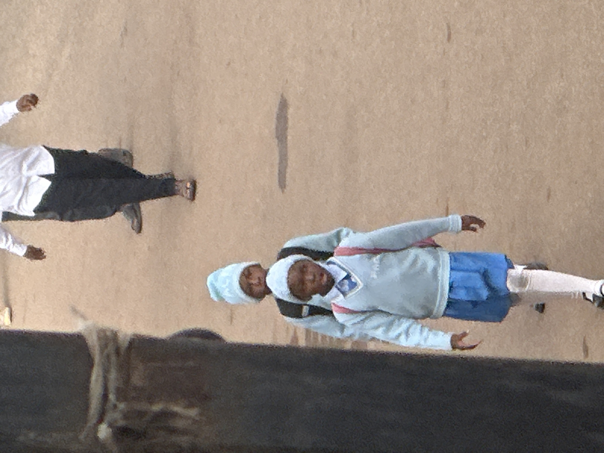 In Bukuru, Nigeria, a group of children in light blue uniforms and beanies walks along a sandy, dirt path. A large, dark pole dominates the left foreground, partially obscuring the children. Two children are prominently visible in the mid-ground, one slightly ahead of the other. The leading child is wearing a light blue sweater vest over a collared shirt, a blue skirt, white knee-high socks, and black shoes. A pink backpack is visible on their back. The second child, walking behind, is wearing a similar light blue sweater, a blue beanie, and a backpack. In the background, a person in a white shirt and dark pants is also walking. The ground appears dusty and uneven.