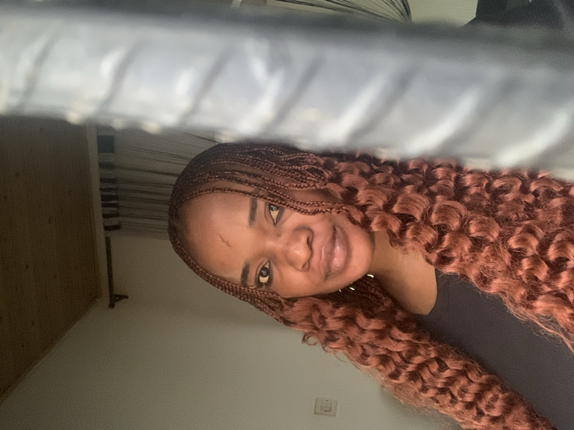 The image is a selfie taken from a low angle of a Black woman. She has brown braided hair with curly ends. She is wearing a dark-colored shirt. The background includes a wall, a door frame, and what appears to be the underside of a bed frame. An electrical outlet can be seen on the wall. The location, based on the user query, is assumed to be Barakin Akawo, Nigeria, although this cannot be verified from the image alone.