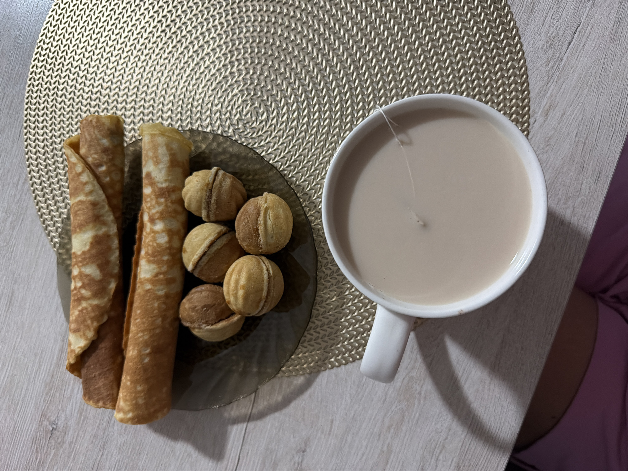 A high-angle shot captures a breakfast setting on a light wooden table, overlaid with a gold woven placemat. In the upper portion of the image, two rolled crepes are stacked, with a small dark plate holding an assortment of six "walnut" shaped cookies arranged in a cluster. Below this, a white ceramic mug filled with a light-colored beverage, likely milk tea or coffee, sits on the placemat. A single white thread is visible suspended over the surface of the liquid within the mug. The scene is illuminated, casting shadows of the objects onto the table. The context of Sirius, Russia, is a detail provided external to the visual information of the image itself.