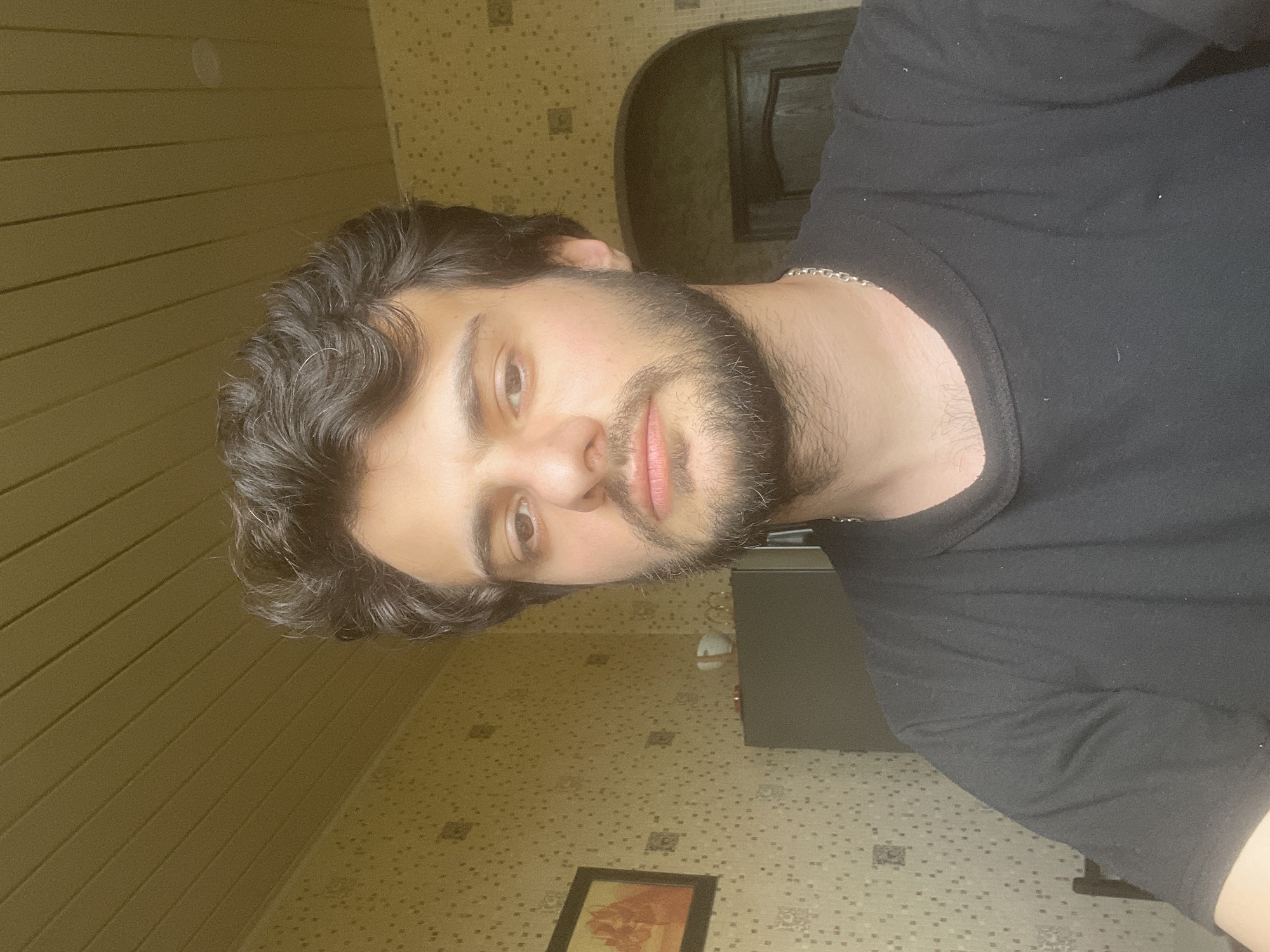 A medium shot is captured of a young man with dark, curly hair and a beard, wearing a black t-shirt and a silver chain necklace. He is looking directly at the camera with a neutral expression. The background features a beige, patterned wallpaper and a wood-paneled ceiling. To the left, a framed picture hangs on the wall. To the right, a dark doorway with an arched top is visible. The overall scene suggests an indoor setting, likely a room in a residential building in Tbilisi, Georgia.