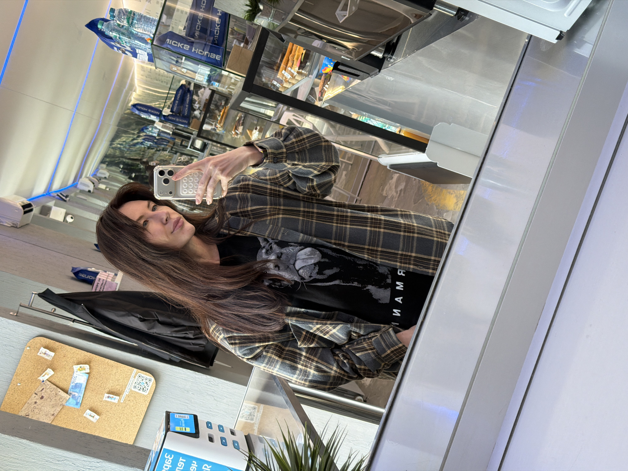 Here is a description of the image.
The image shows a woman taking a selfie in a mirror. She is wearing a black t-shirt with the word "МИАМЯ" on it, and a grey and brown plaid shirt over the t-shirt. She is holding a phone with her right hand, and the reflection of the phone is visible in the mirror. Her hair is long and brown.
In the background, there is a display case filled with drinks and snacks. Above the display case are the words "MICKE HONBE" written on a sign. Also visible in the background is a corkboard with various papers and stickers attached to it. There is also an umbrella hanging above the corkboard. A small potted plant is near the mirror in the foreground.
The name of the city "Perm" and country "Russia" are not identifiable in this image.
