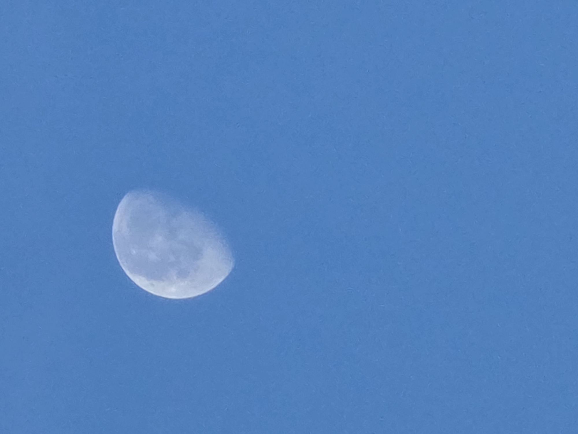 The image shows a close-up view of the moon against a clear blue sky. The moon is partially illuminated, showing a crescent shape. The surface of the moon appears textured, with some visible craters and maria. There are no other objects or people visible in the image. The environment is a sky, indicating an outdoor setting. The image is taken from Kelsterbach, Germany.
