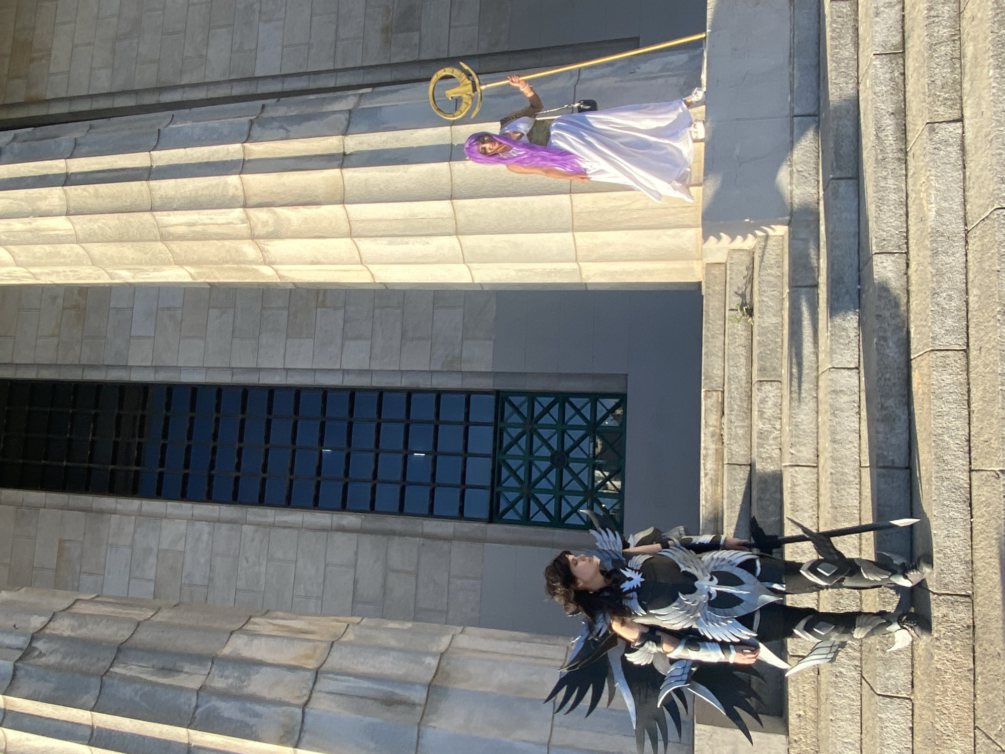 The image shows two individuals in costume posing on a stone structure. One person is dressed in white with purple hair, holding a golden staff. The other is wearing a black and silver outfit with wings. The location appears to be in Buenos Aires, Argentina, identifiable by the architectural style of the building in the background. The specific site may be a historical building or monument with stone construction.