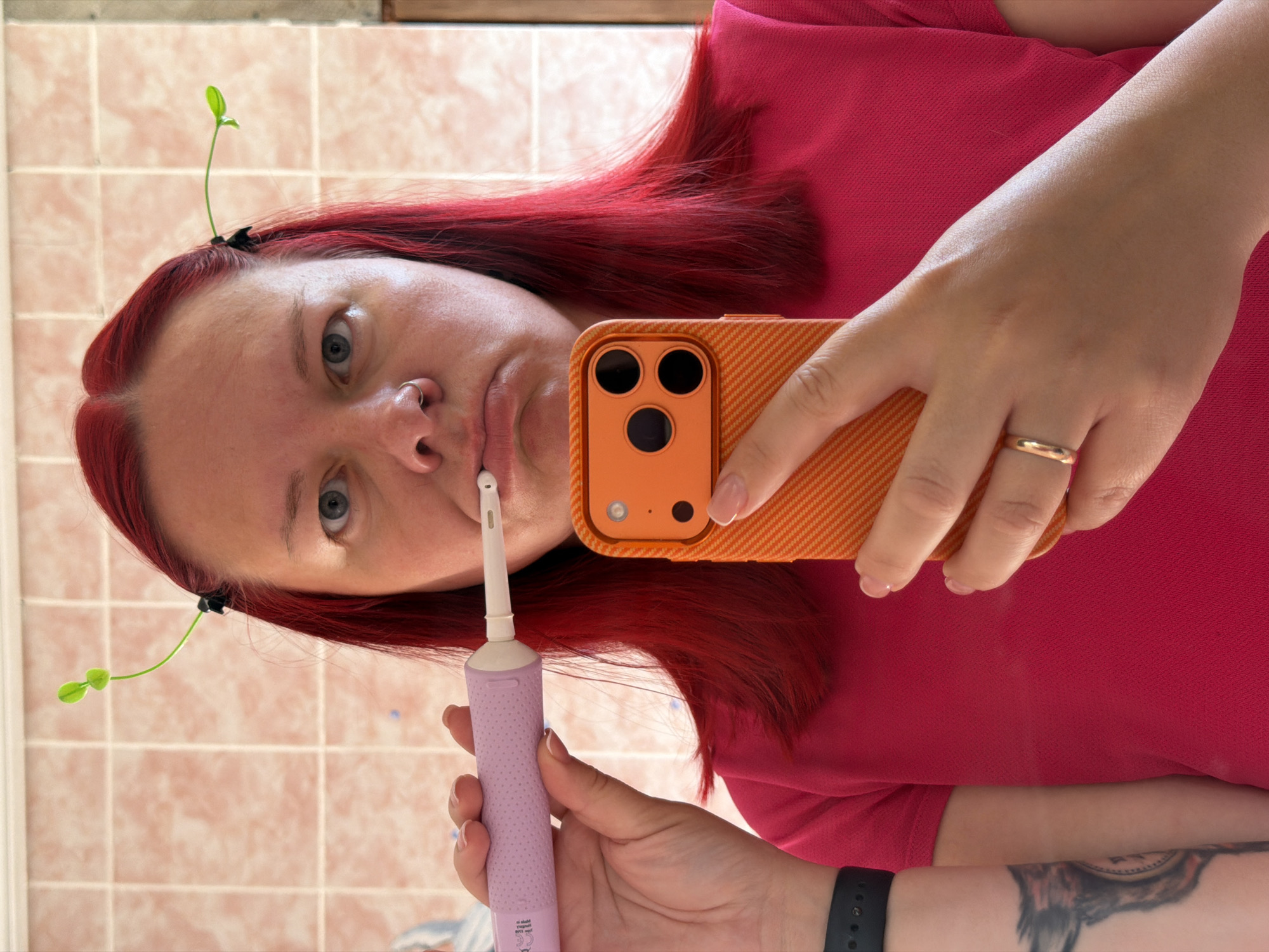 A fair-skinned woman with dyed red hair takes a selfie in a bathroom. The bathroom has light pink square tiles on the wall. She is wearing a red shirt. She is holding an orange smartphone with three camera lenses in her right hand and a light purple electric toothbrush in her left hand, which she has placed in her mouth. There are two green plastic sprouts attached to her hair with black hair clips. A gold ring is visible on her ring finger. A black wristwatch is visible on her left wrist, along with a tattoo. The location of the bathroom is the city of Chaikovsky, Russia.