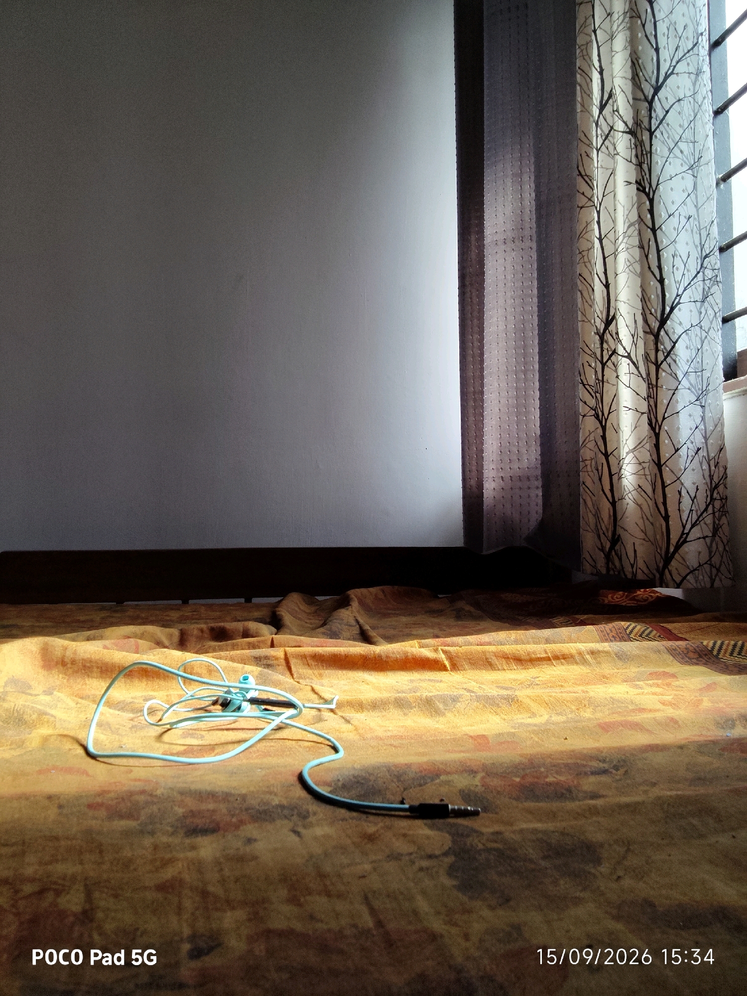 The image depicts an interior scene, possibly a bedroom or living area, with a light-colored wall and a patterned fabric spread across the floor. A pair of light blue earbuds with a black audio jack lies on the fabric. Sunlight streams in from a window on the right side of the frame, partially illuminating the scene and casting shadows. A curtain with a black tree-like pattern hangs near the window. The bottom left corner of the image displays text that reads "POCO Pad 5G," and the bottom right corner shows a timestamp: "15/09/2026 15:34." The location is identified as Kanjany, India. There are no people or animals visible in the image.