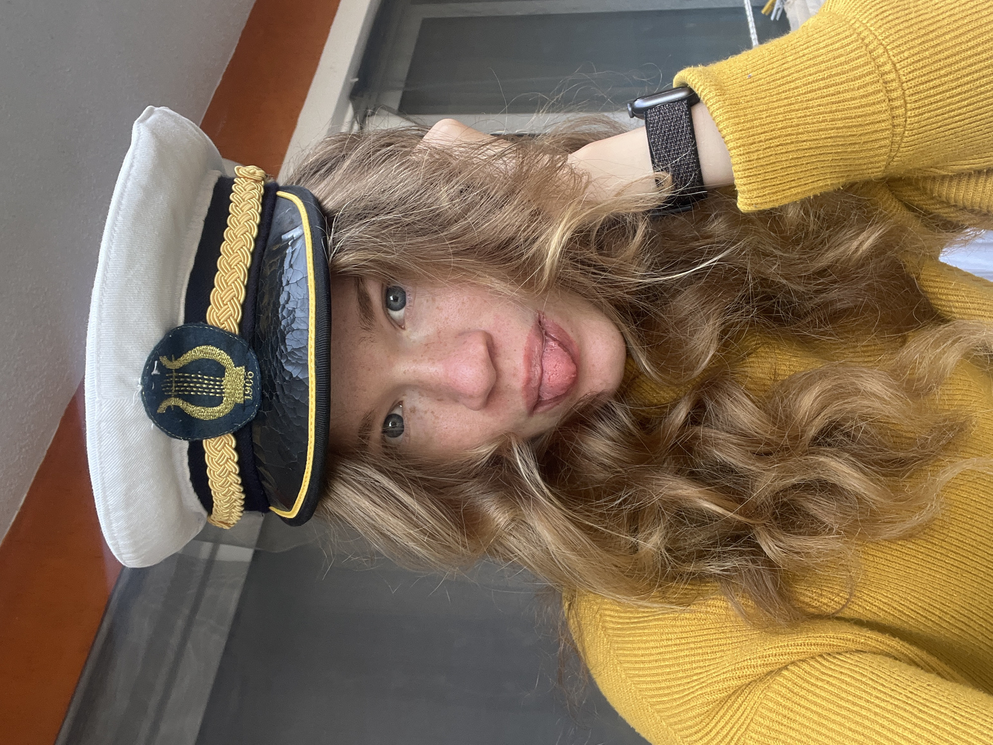 A person with blonde, curly hair, wearing a yellow sweater and a white captain's hat with gold and black accents, is sticking their tongue out at the camera. They are also wearing a black smartwatch on their wrist. The background includes a window with horizontal blinds and what appears to be a wall. The location and city are unknown.