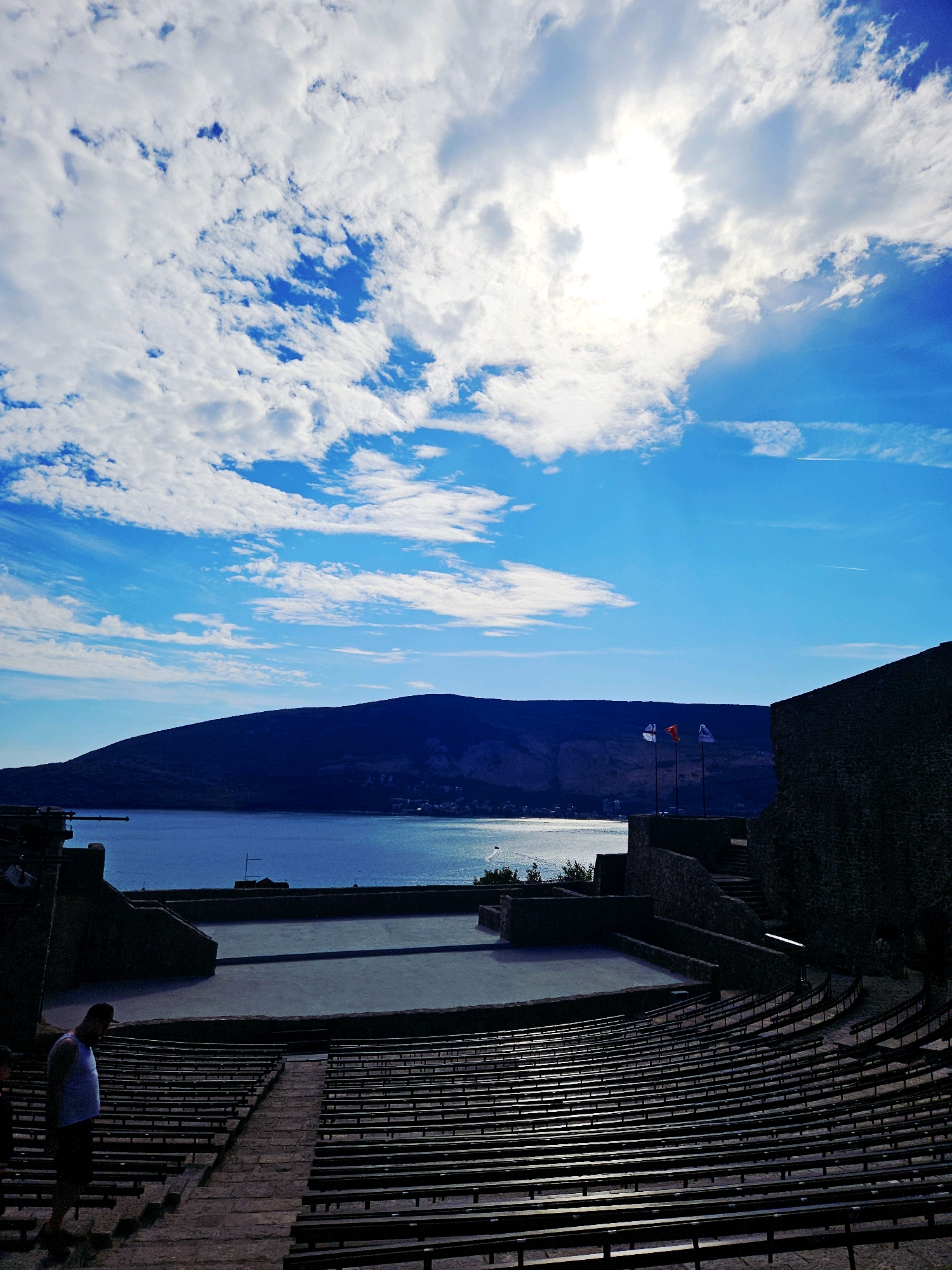 This is a vertical, eye-level, outdoor shot of the Kanli Kula Fortress in Herceg Novi, Montenegro. The fortress, a historic open-air theater, features tiered seating made of dark wood arranged in a semicircular fashion facing a stage area. The stage is a flat, open space. Behind the stage, a stone wall forms part of the fortress structure. Three flags fly from poles on a raised platform near the wall. In the background, a large body of water, likely the Adriatic Sea, stretches out, meeting a dark, mountainous silhouette. The sky above is a vibrant blue, dotted with white, fluffy clouds, and the sun shines brightly through a gap in the clouds, casting light on the scene. To the left of the seating area, a person in a white tank top and dark shorts walks down stone steps. The overall scene depicts a dramatic and scenic location, possibly during daylight hours given the lighting conditions.