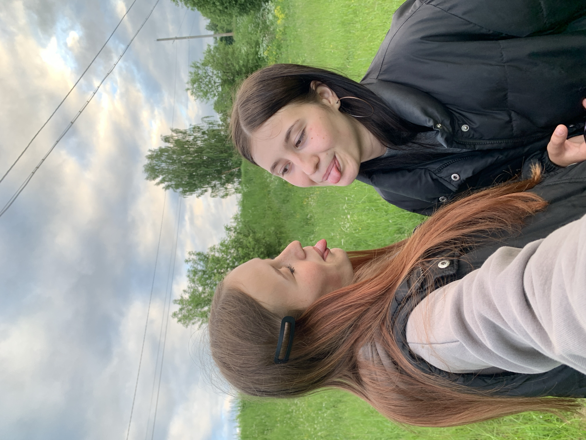 The image depicts two young women outdoors in Фоки, Russia, engaged in a playful interaction. Both subjects are looking towards each other with their tongues extended.
The woman on the left is positioned in profile, facing right. She has long, reddish-brown hair, secured on the right side of her head (viewer's left) with a black rectangular barrette. She appears to have freckles on her nose and cheeks. Her attire consists of a light grey long-sleeved shirt worn under a black sleeveless puffer vest.
The woman on the right is shown in a three-quarter profile, facing left. She has long, dark brown hair and wears a small gold hoop earring in her visible ear. She also displays freckles on her face. Her clothing includes a grey top beneath a black full-sleeved puffer jacket, which is zipped and snapped up to her neck. Her left hand (viewer's right) is visible near the bottom of her jacket, possibly holding a small object.
The background features a vibrant green grassy field, containing scattered white and yellow wildflowers. Behind the subjects, a gentle grassy slope or hill is visible, populated by several deciduous trees with lush green foliage. The sky above is overcast with a mixture of grey and white clouds, with some lighter patches suggesting diffused sunlight. Two parallel utility wires run horizontally across the upper portion of the frame, and a wooden utility pole is partially visible on the far right side of the image.