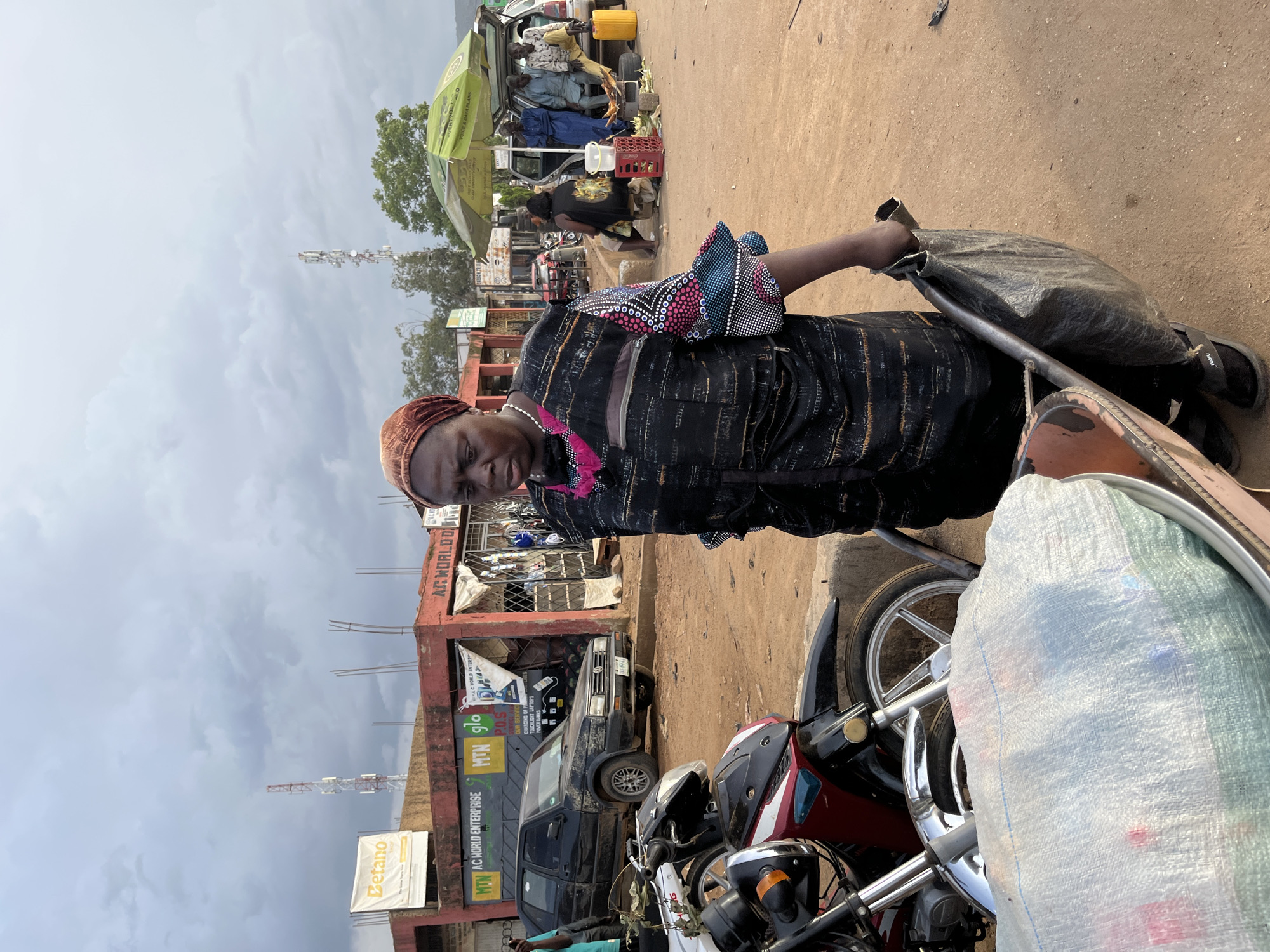 This media file captures a scene in Rafin Par, Nigeria. A woman wearing a traditional dark dress with a patterned yoke and a headscarf stands in the foreground, looking towards the camera. She is holding a large, dark bag in her left hand, and her right arm is bent, with her hand resting on a metal bar. To her right, parts of a motorcycle and a large white sack are visible.
The background depicts a bustling outdoor market. Several motorcycles are parked, and a dark-colored car is stopped in front of a building with shops. The building has signage, including "Betano," "MTN AC WORLD ENTERPRISE," and "AC WORLD ENTERPRISE." Other signs with text like "P.O.S" and "OUR SERVICES" are also visible. Several people are present in the background, some tending to stalls or interacting near the vehicles. A green umbrella shades a portion of the market. In the distance, a telecommunications tower stands against a cloudy sky, suggesting it might be late afternoon or evening. The ground is a dusty, reddish-brown, typical of many Nigerian roads. The overall atmosphere appears to be that of a busy market day.