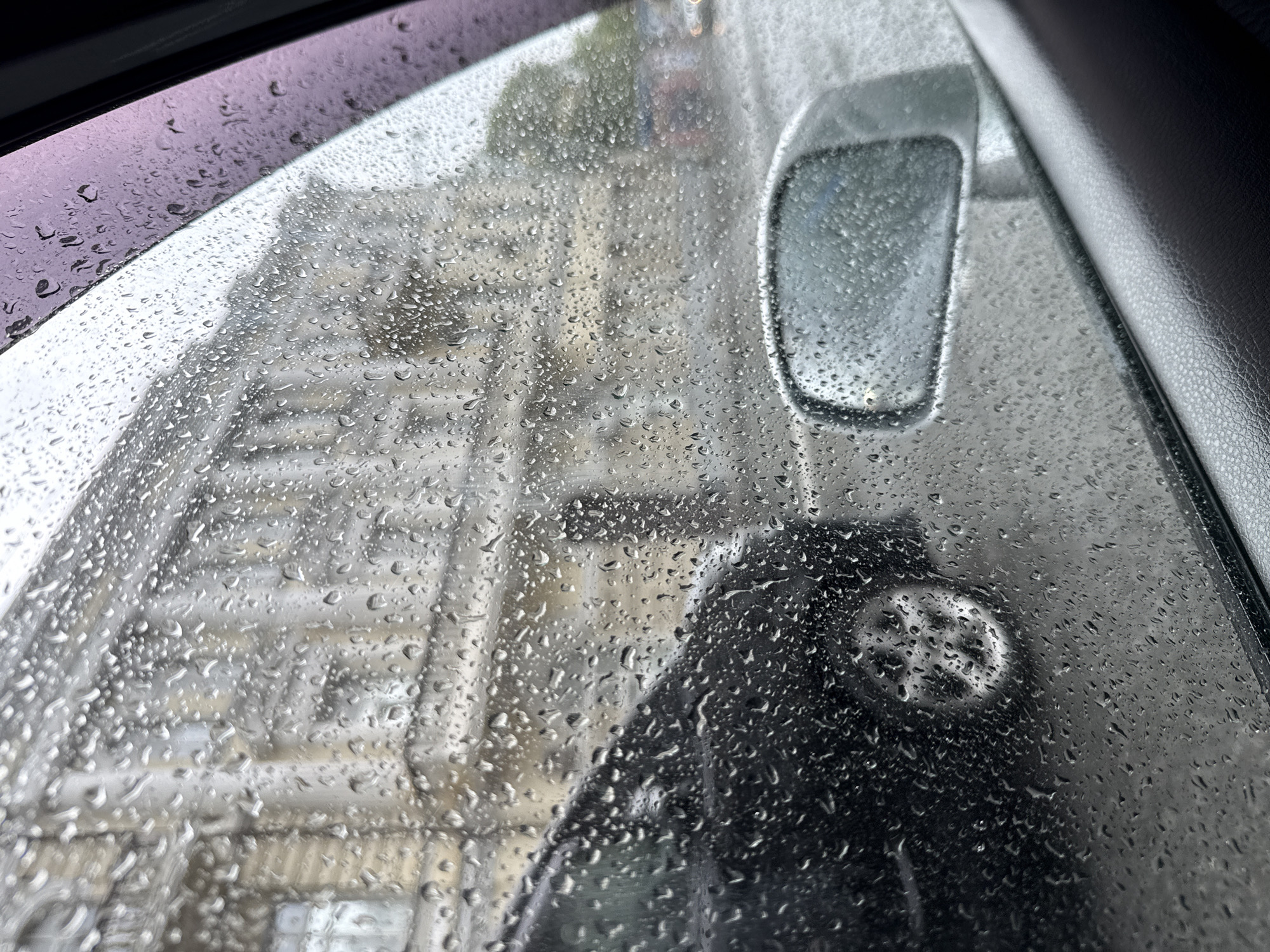 The image captures a close-up view from inside a car, looking out through a side window heavily covered in countless small raindrops. The water droplets distort and blur the exterior scene, creating an impressionistic effect.
Beyond the rain-streaked glass, a cityscape is vaguely visible in the background. Light-colored, multi-story buildings with numerous windows appear as soft, out-of-focus forms, suggesting an urban environment. In the mid-ground, partially visible through the water droplets, is another vehicle. Its side-view mirror is clearly identifiable as a reflective oval shape, and below it, a dark-colored car body is seen, with what appears to be a wheel or hubcap featuring a distinct spoke-like pattern.
Elements of the car's interior frame the view. On the upper left, a dark, possibly purple-tinted plastic or glass surface, likely a window deflector or part of the window frame, is visible and also dotted with raindrops. On the right side, a dark, textured surface, consistent with a car's door panel or interior trim, runs vertically along the edge of the frame.
The weather is clearly rainy, indicated by the widespread water droplets. The lighting is diffused and soft, suggesting an overcast day. There are no people visible in the image, and no discernible text. The scene conveys a sense of enclosure and observation from within a vehicle during a rain shower.