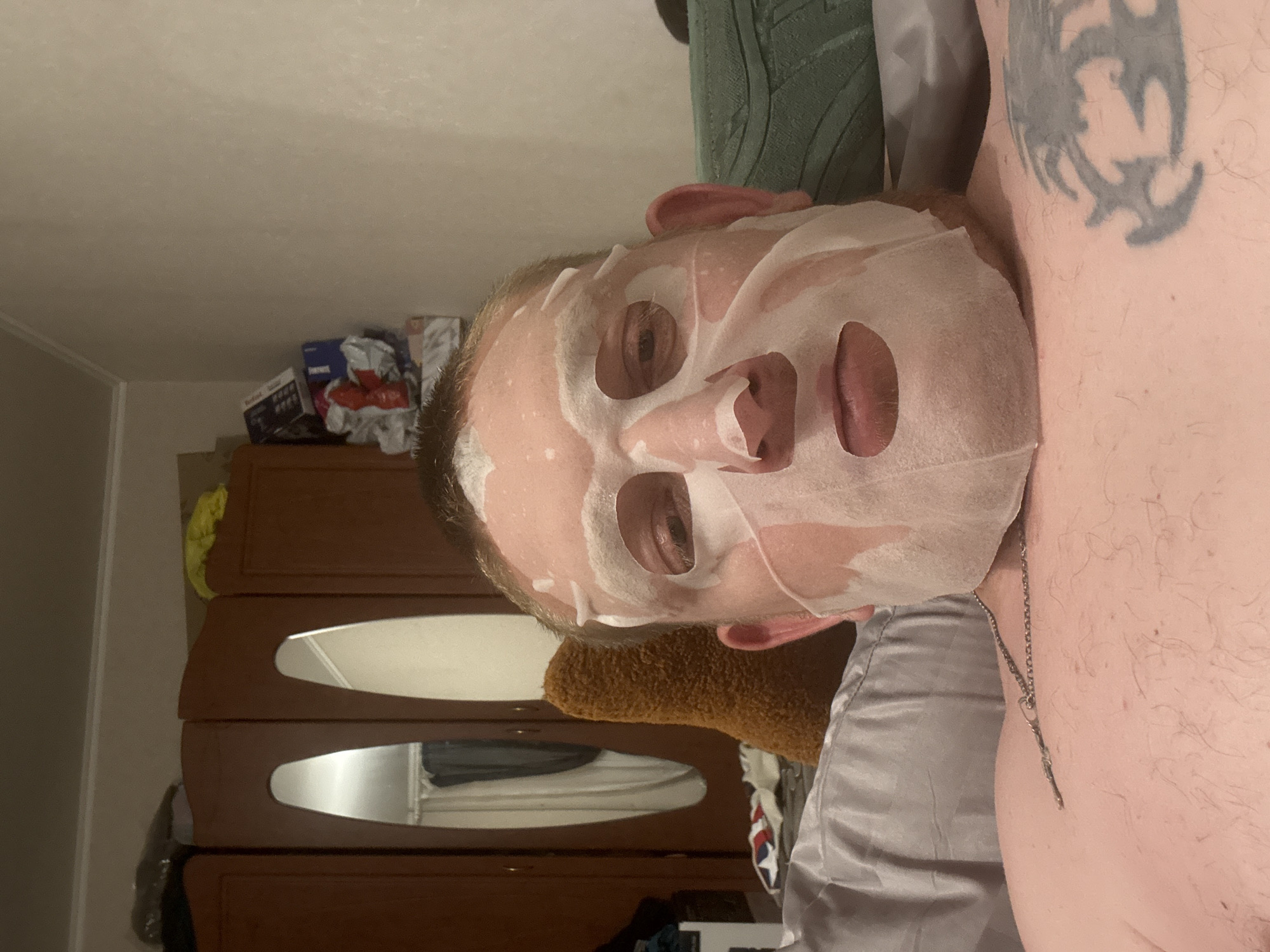 A man is lying on a bed, wearing a sheet face mask. He has a tattoo on his chest and is wearing a necklace. The room appears to be a bedroom, with a wardrobe and other personal belongings visible in the background. The lighting is dim, suggesting it is either nighttime or the room is not well-lit.