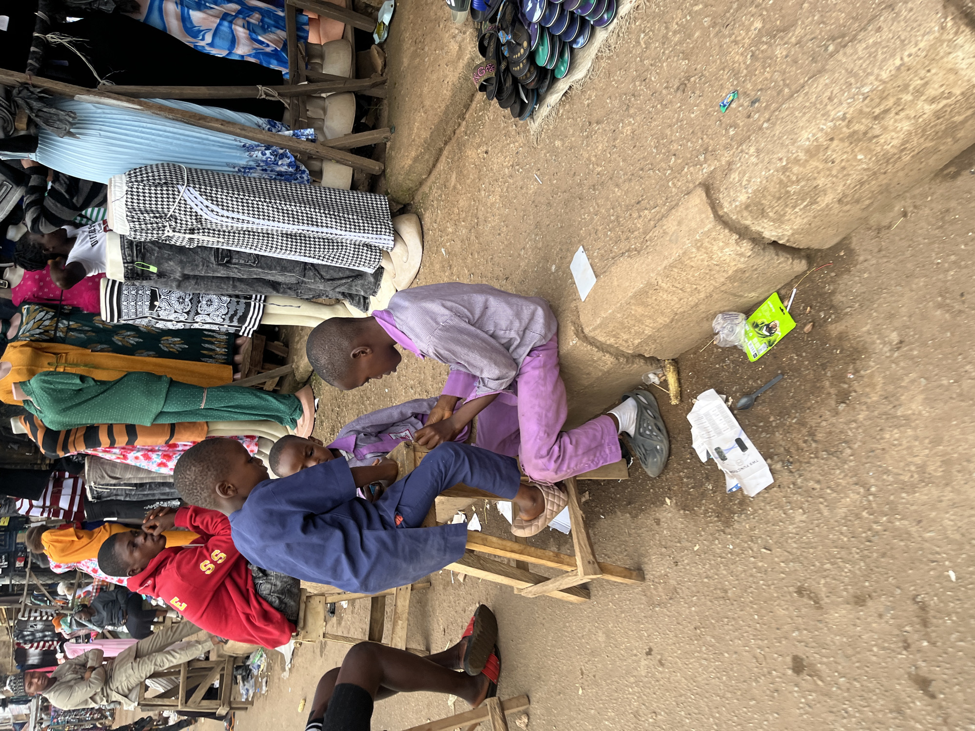 In the city of Bukuru, Nigeria, a street market scene unfolds, captured from a slightly elevated perspective. The foreground is dominated by a dusty, uneven ground, with a raised concrete curb running diagonally across the image, showing signs of wear and a central crack. Scattered on the ground are various pieces of litter, including crumpled paper, a plastic bag, a spoon, and a bright green packet.
Three young boys are the central focus. One boy, wearing a blue tunic and trousers, sits on a rustic wooden stool, bending over and seemingly interacting with another boy. This second boy, dressed in a purple checkered shirt and purple trousers, is also seated on a wooden stool and is actively engaged in something small held between his fingers, possibly a task or game. A third, even younger boy, is partially visible between the two older boys, wearing a purple shirt and looking towards the ground.
To the left, a fourth boy, partially cropped, sits on the ground with his legs extended, wearing black shorts and red sandals. His posture suggests he is resting or observing.
In the background, the market bustles with activity, though many figures are blurred and indistinct. Mannequins dressed in brightly colored clothing, including green, yellow, and blue dresses, are displayed on wooden stands, suggesting a clothing stall. Various fabrics and garments are visible on racks and tables, hinting at the variety of goods for sale. A man in a light-colored traditional outfit sits on a high wooden stool on the far left, observing the scene. The overall impression is one of a vibrant, everyday market environment.
