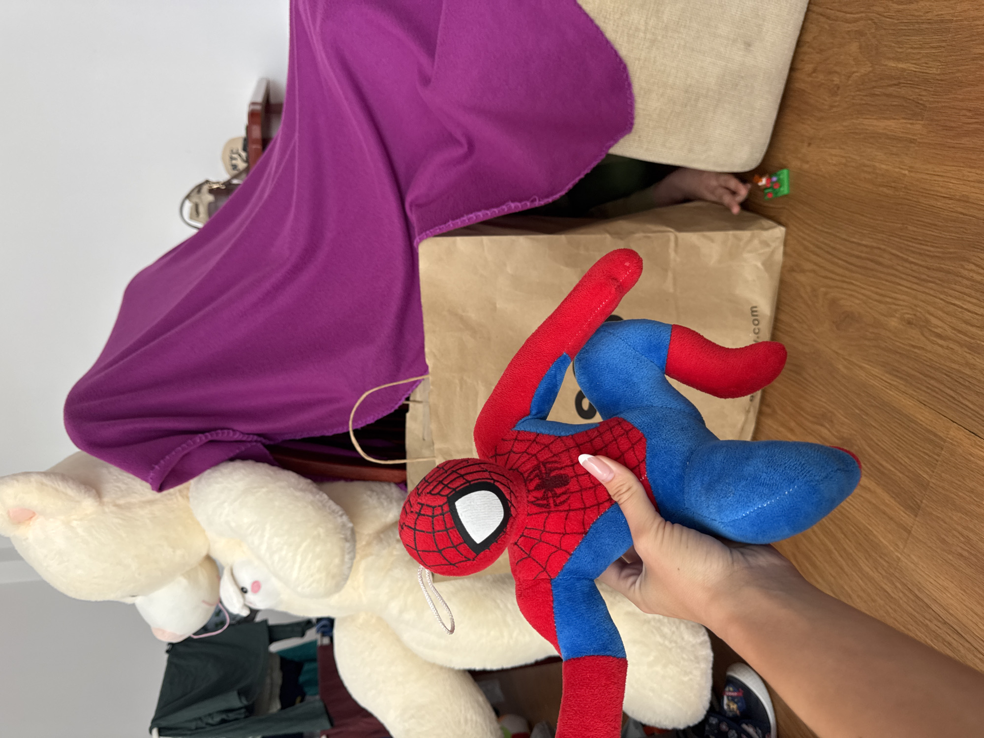 A person holds a stuffed Spiderman toy. A large white teddy bear, a purple blanket, and a brown paper bag are in the background. A child's hand and a small toy are visible to the right of the bag. The location is not specified, and there is no way to tell if it is Burgas, Bulgaria.