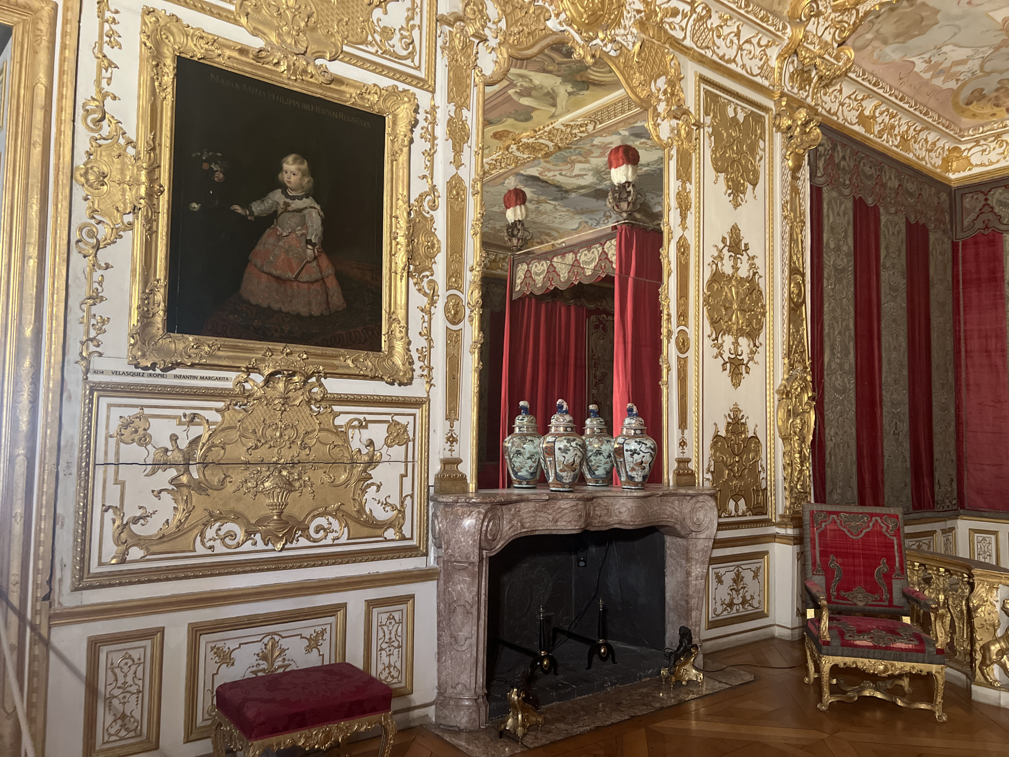The image depicts an opulent interior of a room, likely a palace or grand residence, in Oberschleißheim, Germany. The room is adorned with intricate golden ornamentation on the walls and ceiling, reflecting a Baroque or Rococo style. To the left, a large framed painting hangs on the wall, depicting a young child in elaborate attire. In the center of the room, a marble fireplace with ornate carvings serves as a focal point, topped with a row of decorative ceramic vases. Red velvet drapes frame a bed or alcove in the background, visible in a mirror that reflects the opposite side of the room and the ceiling's painted frescoes. To the right, a richly upholstered red armchair with golden accents sits on the polished wooden floor, adjacent to a gilded railing. The overall impression is one of extreme luxury and historical significance.