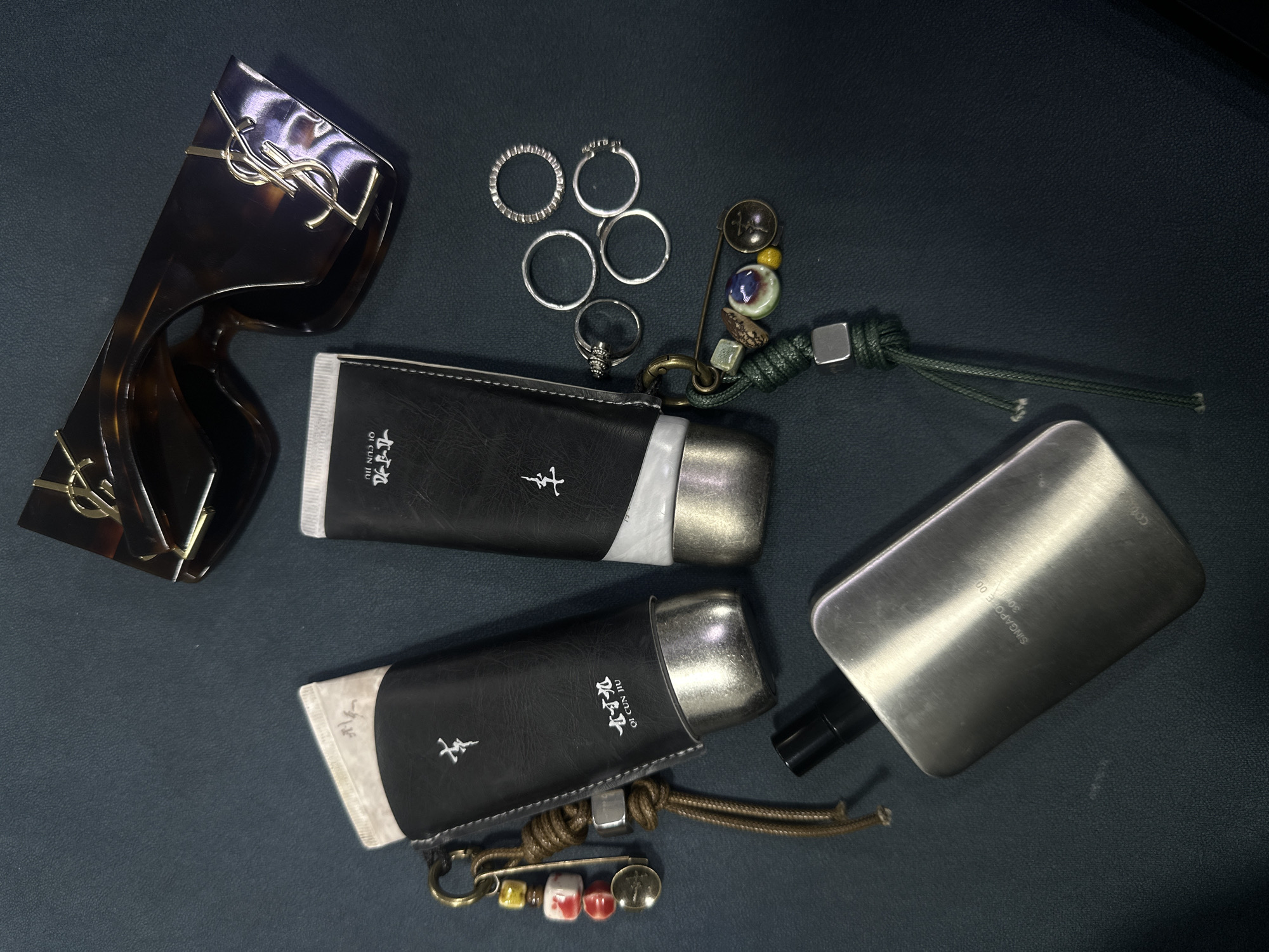 The image shows a dark surface with various personal items scattered across it. There are brown tortoiseshell sunglasses with a prominent gold YSL logo in the upper left corner. To the right of the sunglasses are several silver rings clustered together. There are also two cylindrical objects encased in black leather holders with white trim, each adorned with a small symbol and Chinese characters. A silver rectangular container with a black nozzle is situated on the right side of the image. A decorative safety pin with beads, a vintage button, and a green knotted cord is present near the rings and the top cylindrical object, while another similar safety pin with beads is attached to the bottom cylindrical object with a brown cord. The location is not discernible from the objects in the image. Yerevan, Armenia, cannot be verified based on the visible content.