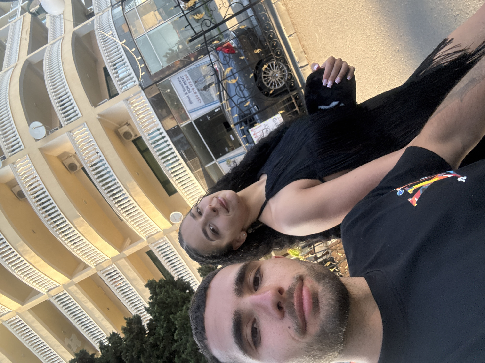 A young couple is posing for a selfie on a street in Nesebar, Bulgaria. The man is in the foreground, looking directly at the camera. He has short dark hair, a beard, and is wearing a black t-shirt with a colorful graphic on the chest. The woman is standing behind him to the right, also looking towards the camera with a slight smile. She has long dark curly hair and is wearing a black fringed dress.
Behind them, a multi-story building with curved balconies and white railings dominates the background. The building appears to be a hotel or apartment complex. There is also a dark-colored car parked in front of a storefront. A sign on the storefront reads "Bikes & Scooters FOR RENT". The sun is shining, casting a warm glow on the scene. The overall impression is a casual moment captured during a vacation.