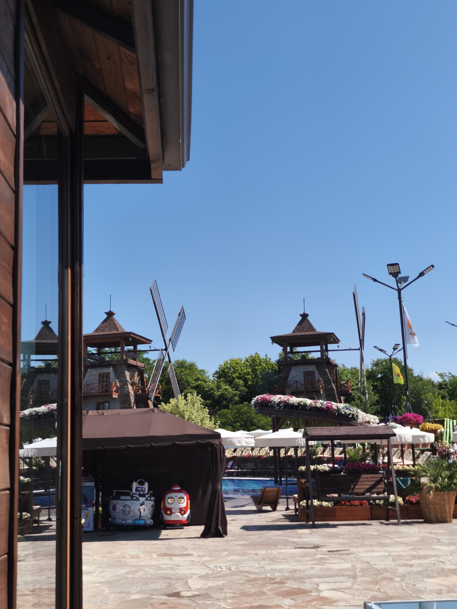 The image shows an outdoor scene with several windmills in the background. In the foreground, there is a brown tent with two toy rides underneath. To the left of the tent, a glass window reflects the windmills. Further back, a swimming pool area is visible with lounge chairs and umbrellas. Trees and a lamppost with flags are also present in the background. The ground is paved with large, flat stones. The sky is clear and blue.
