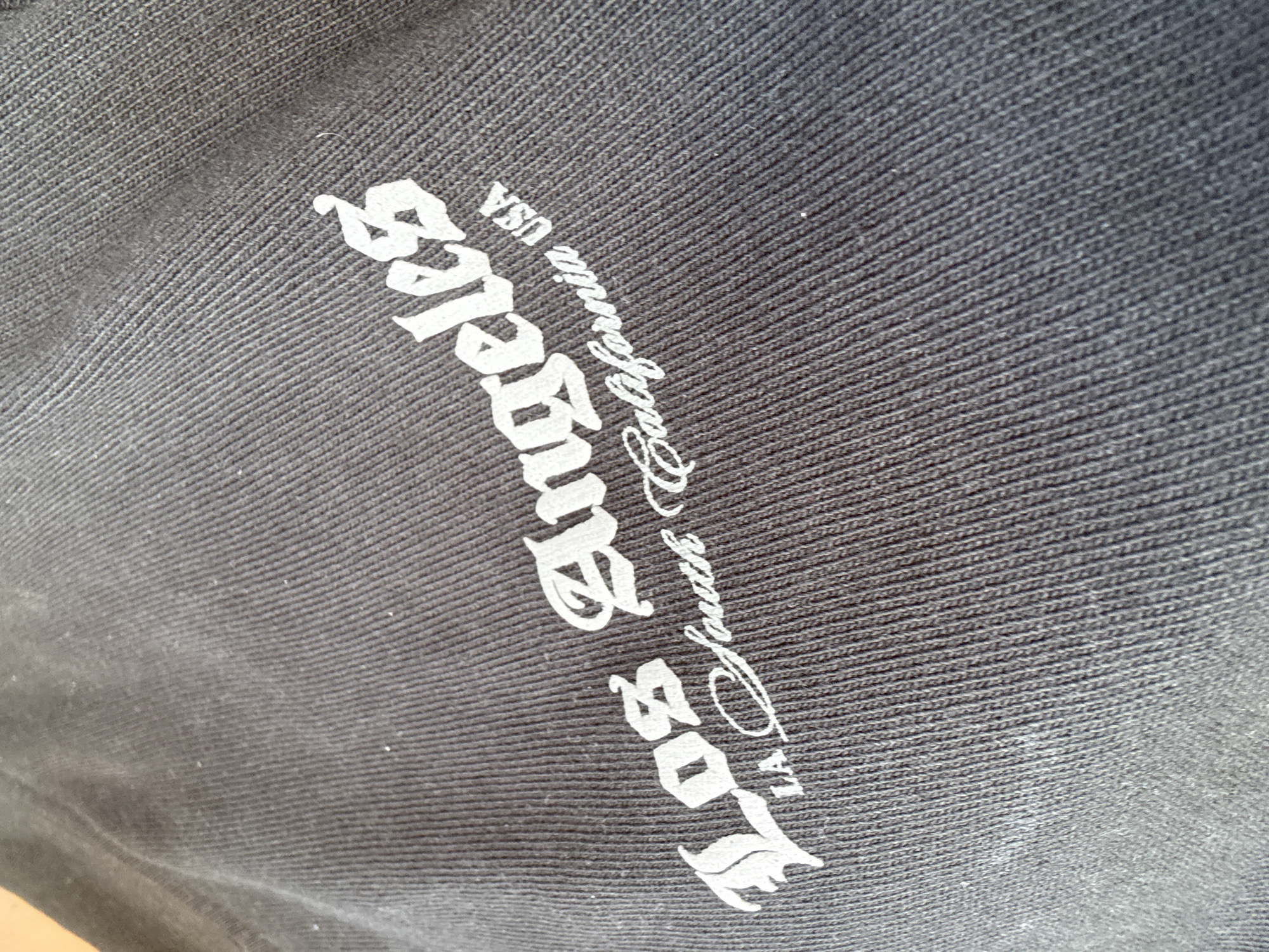 The image displays a close-up, angled view of a dark, ribbed fabric, likely a garment. Emblazoned on the fabric in white, stylized lettering are the words "Los Angeles" in a gothic font, followed by "LA South California USA" in a script font below it. The fabric appears to be a t-shirt or sweatshirt, and the print is the primary subject. There are no visible people, actions, or interactions. The environment is undefined beyond the texture and color of the fabric. There is no information in the image to suggest a location in Draginovo, Bulgaria.