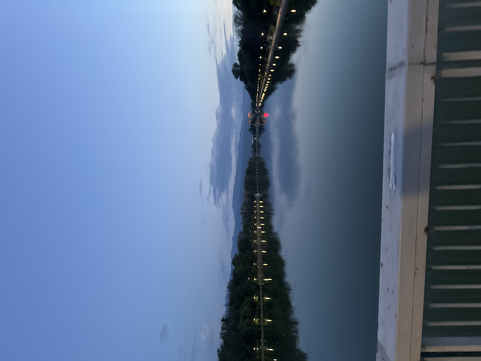 The image, when correctly oriented, captures a serene twilight scene over a wide body of water, likely a canal or a river, framed by an illuminated promenade and distant mountains under a cloudy sky. The sky, a deep blue, transitions from a darker tone at the zenith to a lighter hue near the horizon, dotted with soft, indistinct clouds.
Along the far bank of the water, a dense line of trees forms a dark silhouette, fronted by a long, continuous row of bright artificial lights. These lights, possibly streetlights or decorative lamps, create vivid, elongated reflections that stretch across the calm, dark surface of the water, mirroring the scene above.
In the mid-ground, a distinctive building or structure is visible on the far bank, emitting a striking red light that produces a prominent red reflection on the water. Further in the distance, a range of mountains is faintly silhouetted against the fading light of the horizon.
On the immediate right of the foreground, part of a white and green structure, appearing to be a bridge railing or a pier, is partially visible. No people or specific activities are discernible in the scene. The overall ambiance is peaceful and tranquil, typical of an early evening or dusk setting. Given the context of Plovdiv, Bulgaria, this landscape likely depicts the city's Rowing Canal, known for its length and illuminated banks. No text is visible in the image.