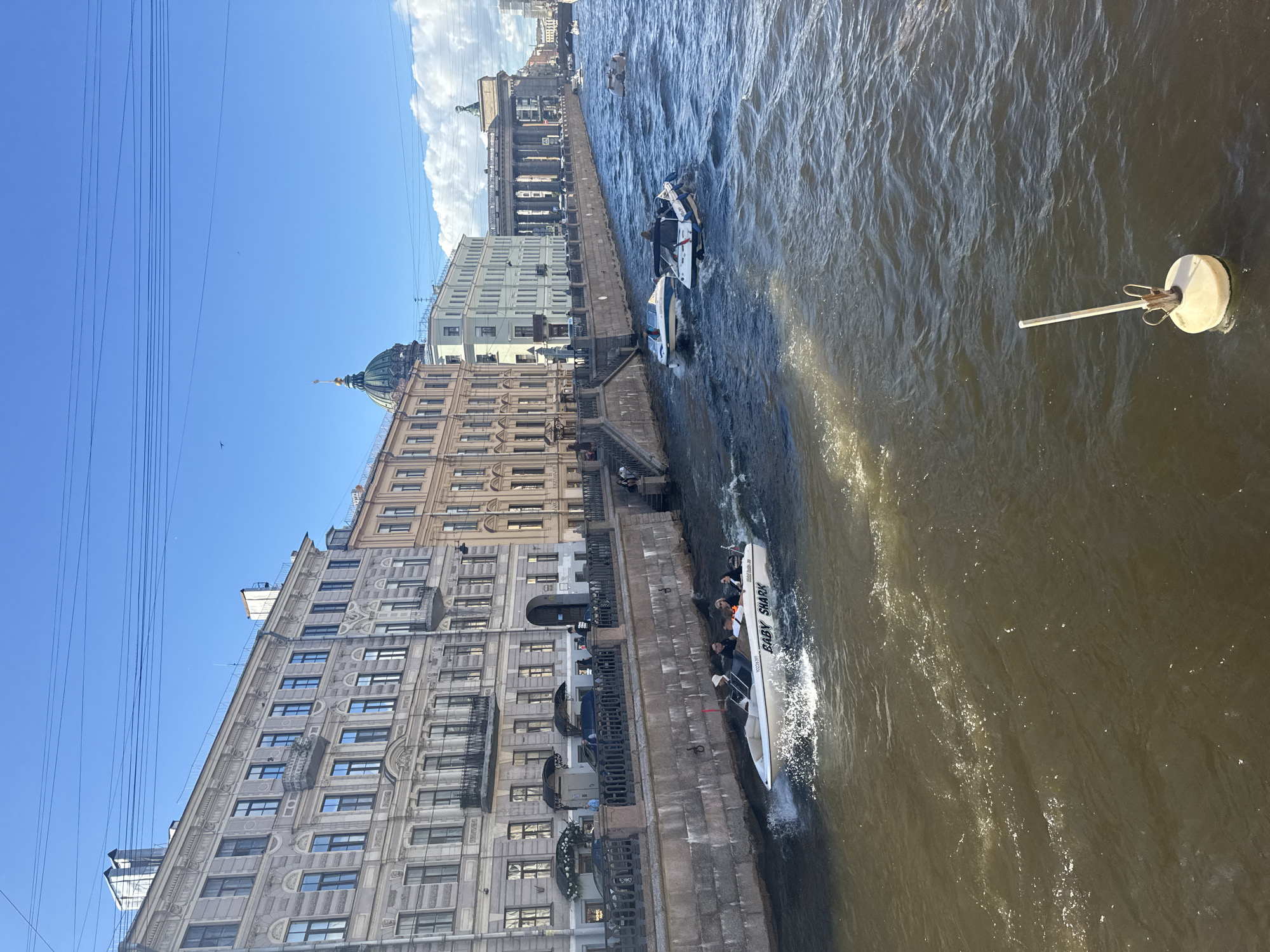 The image captures a vibrant canal scene in Saint Petersburg, Russia, likely along the Griboyedov Canal or Moyka River, given the characteristic architecture and waterway. Several boats are actively navigating the choppy, brown water. In the foreground, a white speed boat with the name "BABY SHARK" emblazoned on its side carries four passengers. Further down the canal, other boats are visible, some with passengers, suggesting a popular tourist or recreational area. The canal is lined with grand, historic buildings featuring classical architectural elements, including ornate facades, balconies, and arched windows. The sky is a clear blue with a few scattered clouds, and overhead, power lines crisscross the frame. A distinct green-domed building, possibly a church, is visible in the background, a common landmark in the city. The overall impression is a lively, bustling urban waterway set against a backdrop of impressive historical architecture.