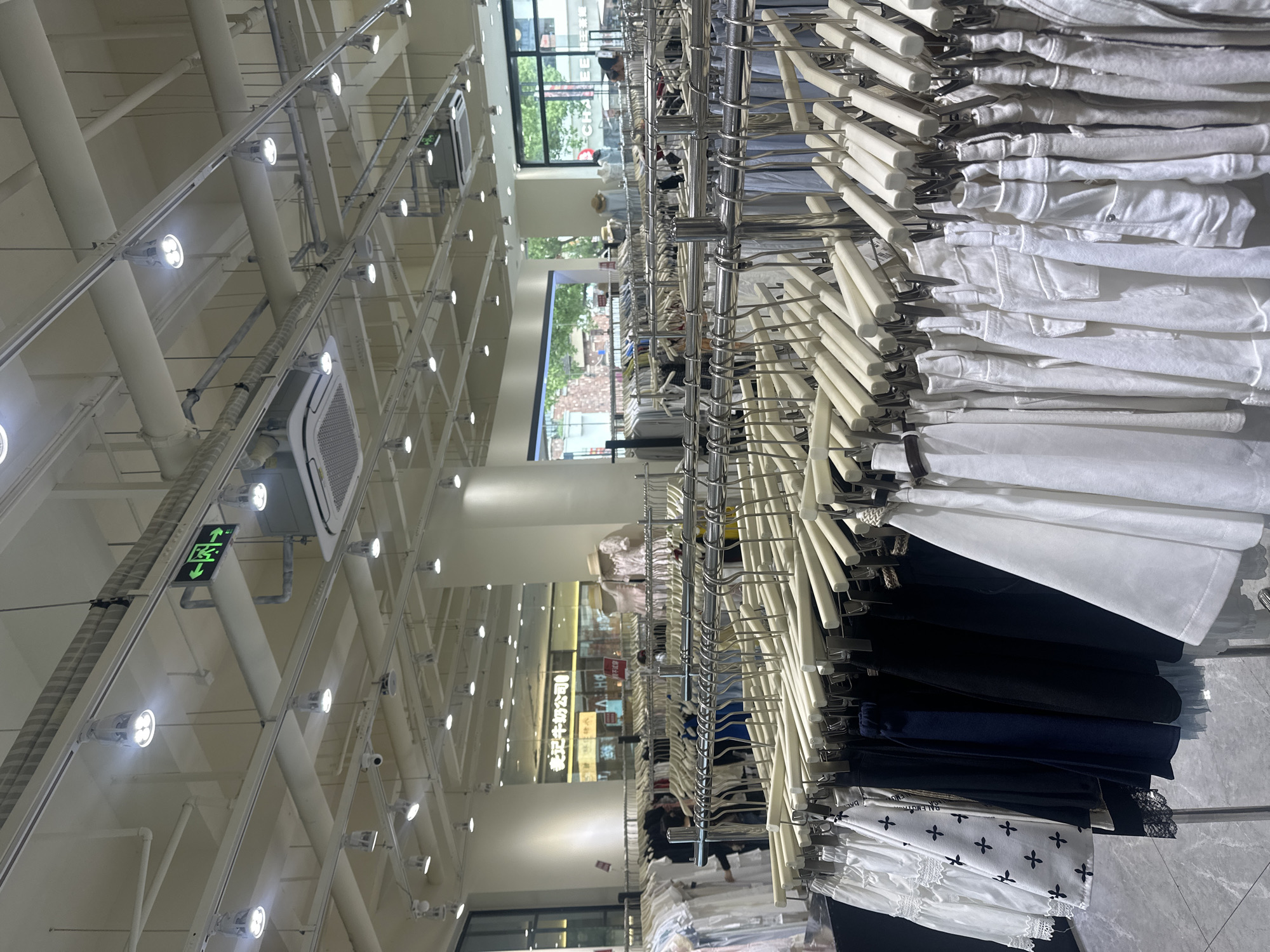 The image shows the interior of a clothing store in Chongqing High-tech Zone, China. Racks filled with clothes on hangers dominate the foreground. The clothing includes white tops and dark-colored bottoms. The store features a high ceiling with a grid pattern of structural supports, lighting fixtures, and air conditioning units. Through windows and store openings, glimpses of other shops and the outdoors are visible. People are seen in the background browsing through the store.