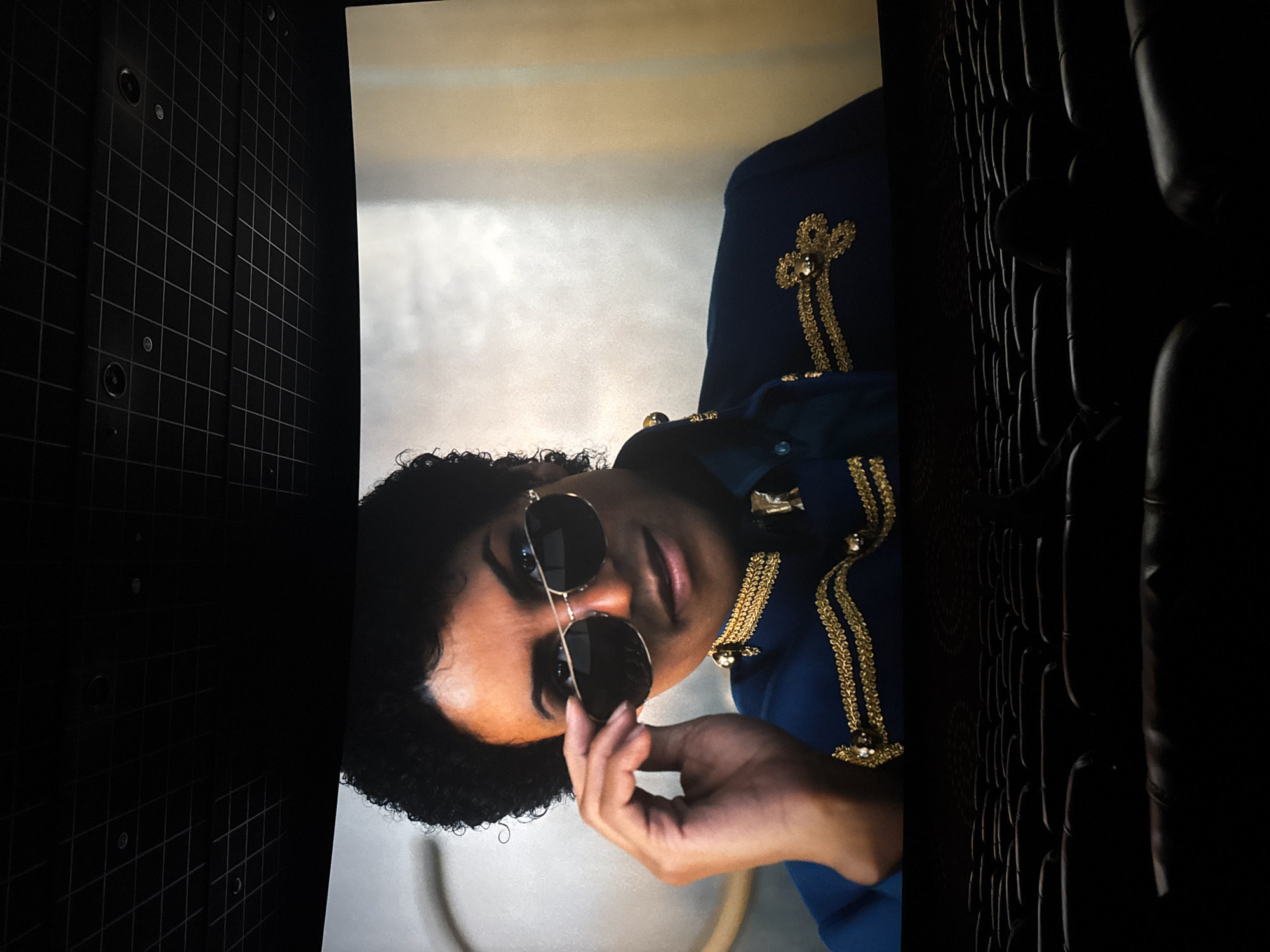 The image is a close-up of a person wearing round sunglasses and a dark blue jacket with gold embellishments. The person has curly hair and is holding the sunglasses. The background behind the person appears to be a screen or display, with a dark grid-like pattern on one side and a pattern of vertical dark ovals on the other.