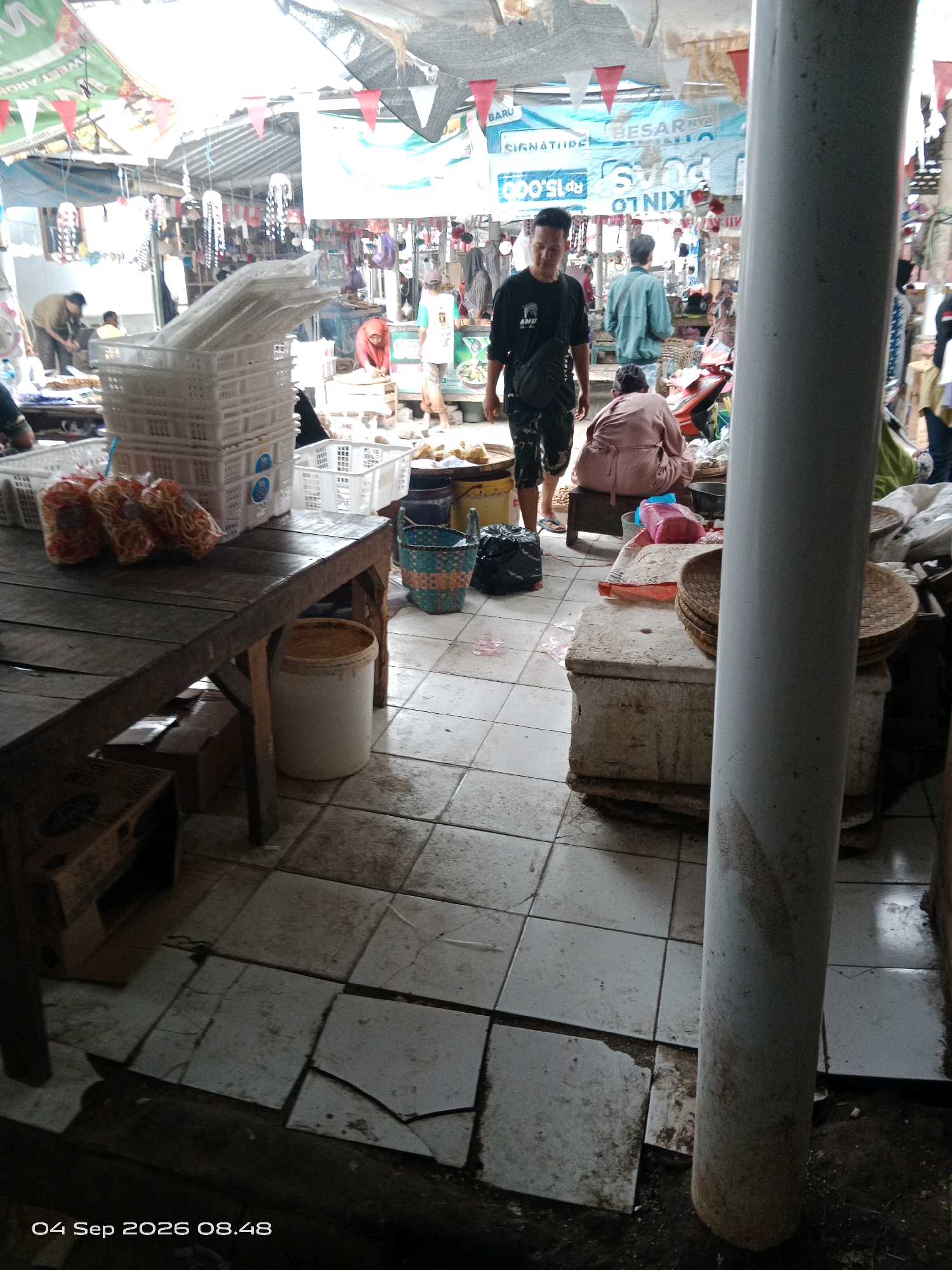 In the city of Kalipurwo, Indonesia, a bustling market scene unfolds. The foreground is dominated by a wooden table laden with white plastic crates and several bags of snacks. To the left, a portion of a cardboard box is visible. A large white cylindrical object, possibly a pole, stands prominently in the right-center of the frame, partially obscuring the background.
The market floor is tiled, with several tiles cracked and displaced, suggesting a weathered environment. Various stalls and vendors occupy the background, with awnings and tarps providing shade. Several people are present, including a man in a black t-shirt and patterned pants walking towards the foreground, a person in a pink garment crouching, and another individual in a green jacket standing further back. Other market-goers are visible in the distance. The overall atmosphere is that of an active, open-air market.