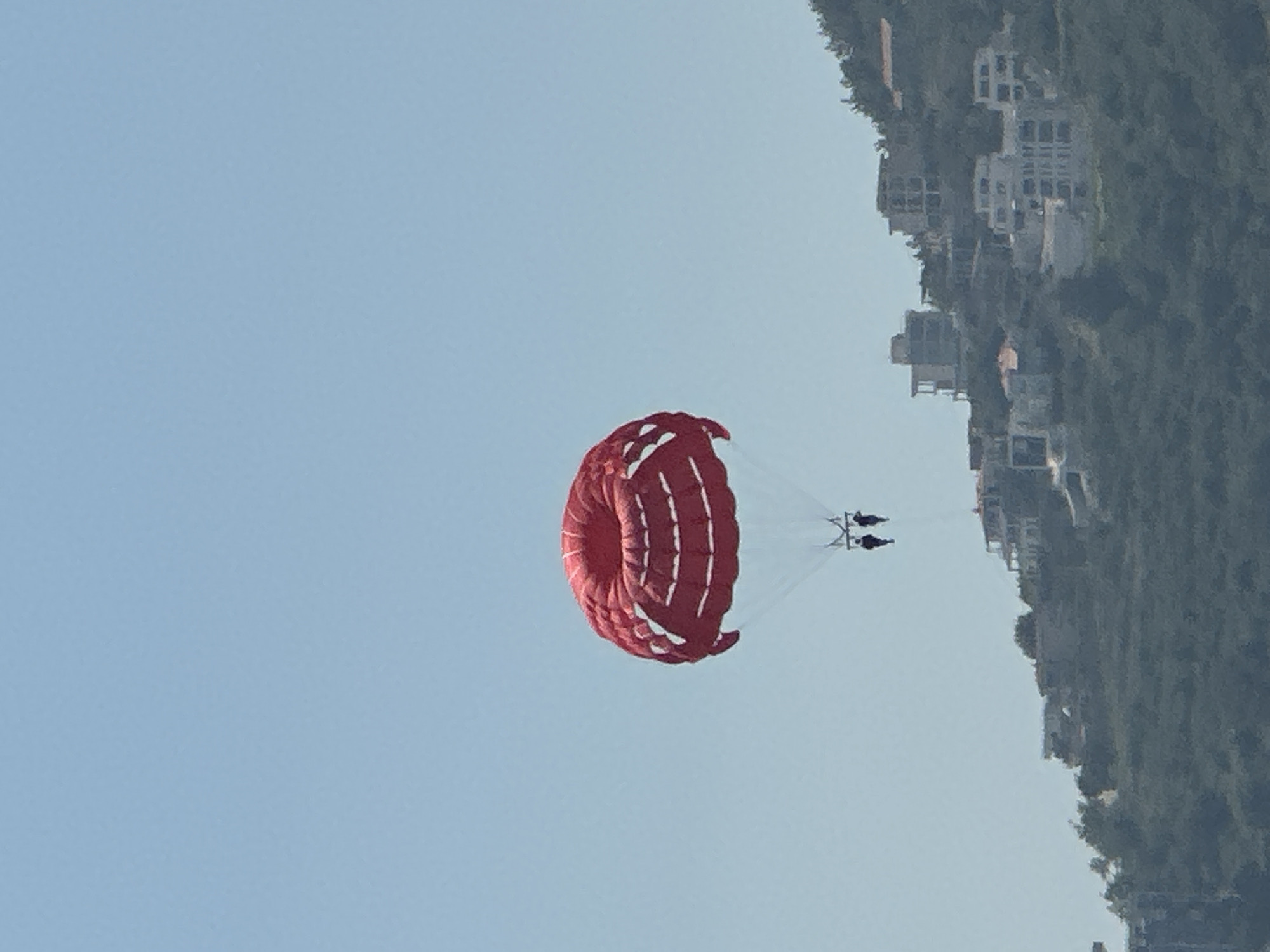 A bright red parasail is suspended in a clear blue sky over the coastal city of Budva, Montenegro. Two people are attached to the parasail, their dark figures silhouetted against the sky. Below the parasail, a lush green hillside is covered with trees and dotted with numerous white buildings, indicative of a developed urban area. The architecture of the buildings suggests a modern Mediterranean style, with multiple stories and balconies. The overall scene captures a recreational activity against a backdrop of a scenic coastal landscape and urban development.