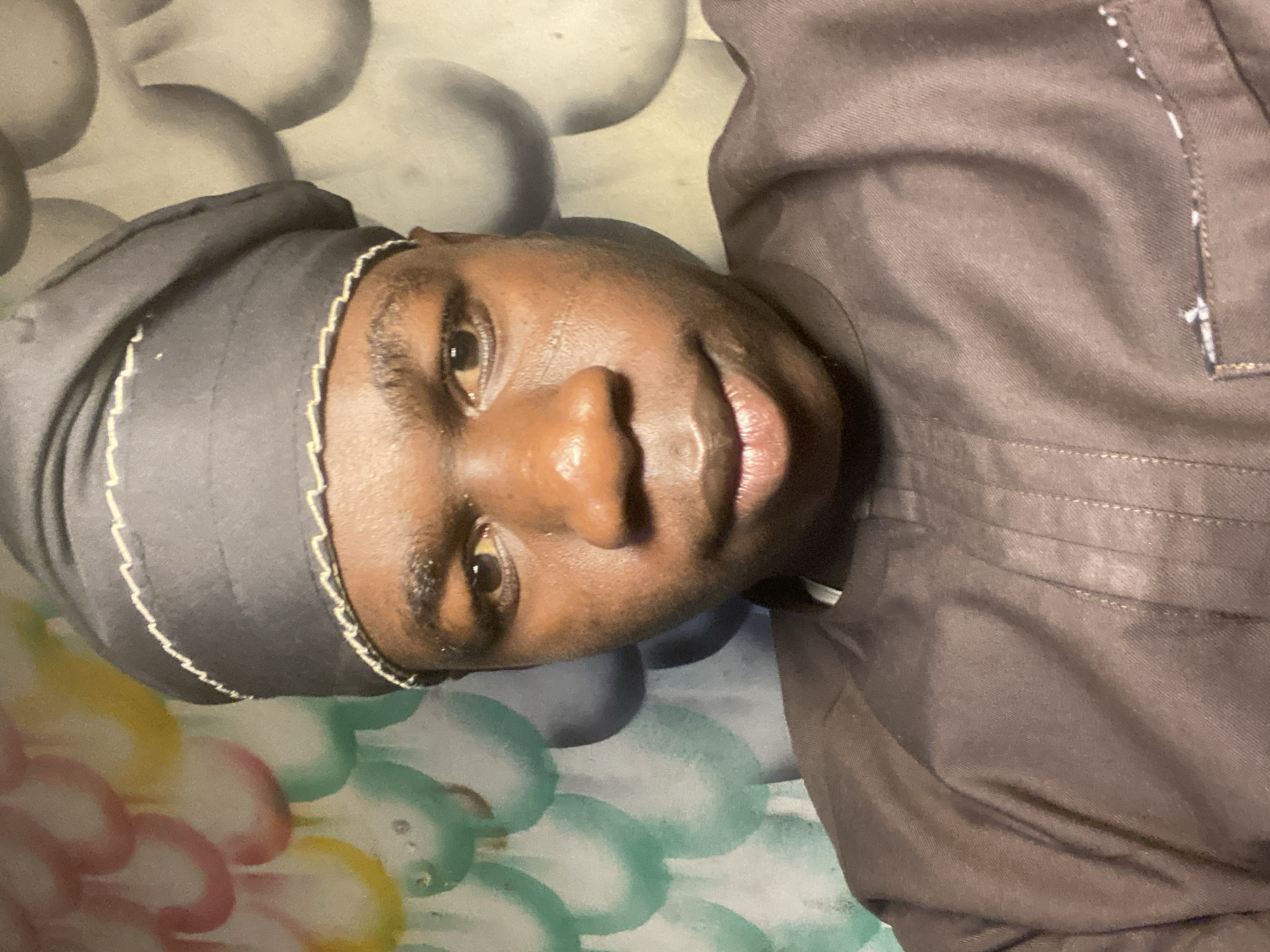 An individual is featured in a close-up, rotated portrait, appearing to lie down in an indoor setting. The subject has a dark complexion, looking directly at the camera with a neutral to slightly soft expression, eyes open and mouth slightly agape. They are wearing a dark grey or brown traditional garment, comprising a head covering with a light-colored, possibly white or cream, stitched or embroidered trim along its edge. A matching dark top with subtle vertical seams or folds is also visible. The background consists of patterned fabric, likely bedding or a cushion. The upper part of the background shows large, soft grey and white abstract shapes, while the lower part displays colorful, irregularly shaped patterns in shades of pink, yellow, green, and light blue. The scene suggests a domestic environment, possibly in Mararaba, Nigeria. The lighting appears to be indoor, and there are no discernible weather conditions or visible text.