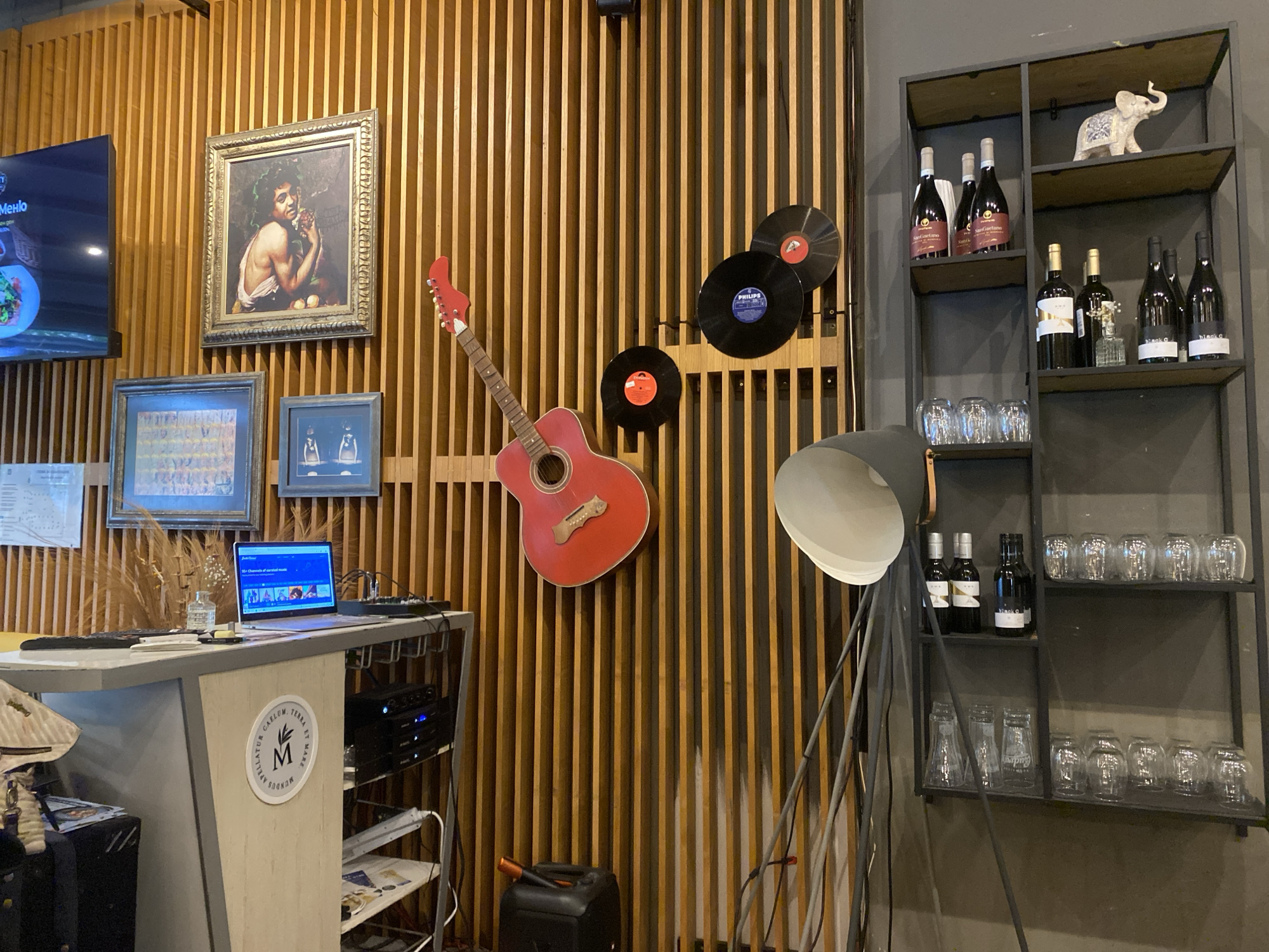 The image captures a restaurant or bar interior, potentially in Varna, Bulgaria, based on the Cyrillic script menu visible on a screen. The space is characterized by a light-colored wooden slat wall adorned with various items: a framed painting of a child with fruit, two framed pictures, a red guitar, and three vinyl records.
To the left is a reception or service desk with a light-colored surface and cabinet. A laptop is on the desk, displaying a screen with various icons and menu options. A black case sits on the floor beside the desk.
To the right of the wooden wall, a dark metal shelf is stocked with wine bottles, glassware, and a small ceramic elephant figurine. A large, grey floor lamp with a broad lampshade stands adjacent to the shelf, casting light toward the center of the room. A black canister is on the floor in front of the wooden wall.
The overall impression is of an eclectic and stylish interior design, combining artistic and musical elements with practical bar or restaurant storage.