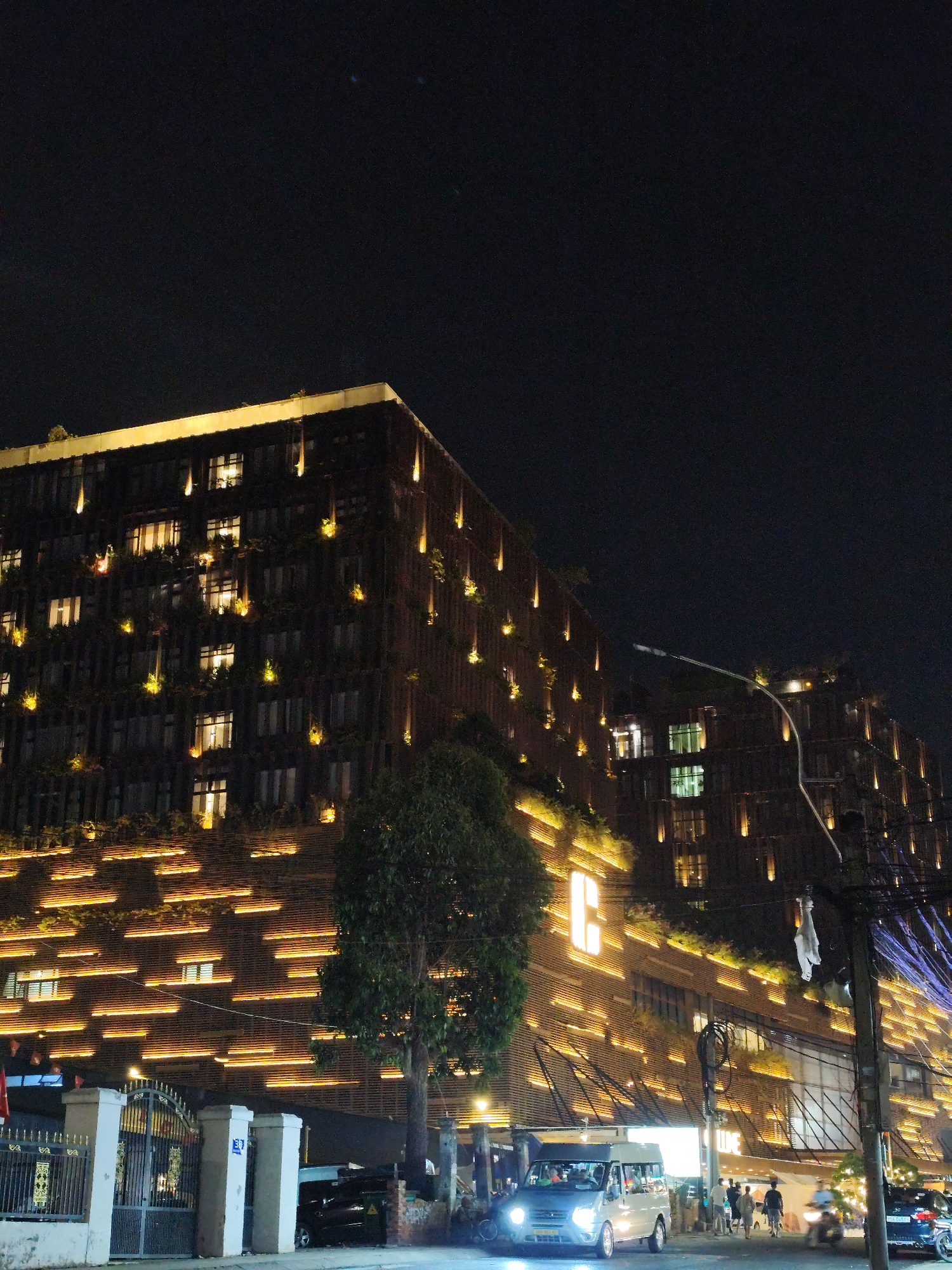 The image depicts a nighttime scene in Da Lat, Vietnam, featuring the illuminated facade of the "C Central Hotel Dalat". The building's exterior is characterized by linear horizontal lighting patterns and vertical accents of greenery. A van is positioned in the foreground, its headlights illuminating the street. Pedestrians and motorcycles are visible on the sidewalk in front of the hotel. The setting includes urban infrastructure like a white fence, utility poles with tangled wires, and streetlights. The overall ambiance is vibrant due to the hotel's elaborate lighting scheme.