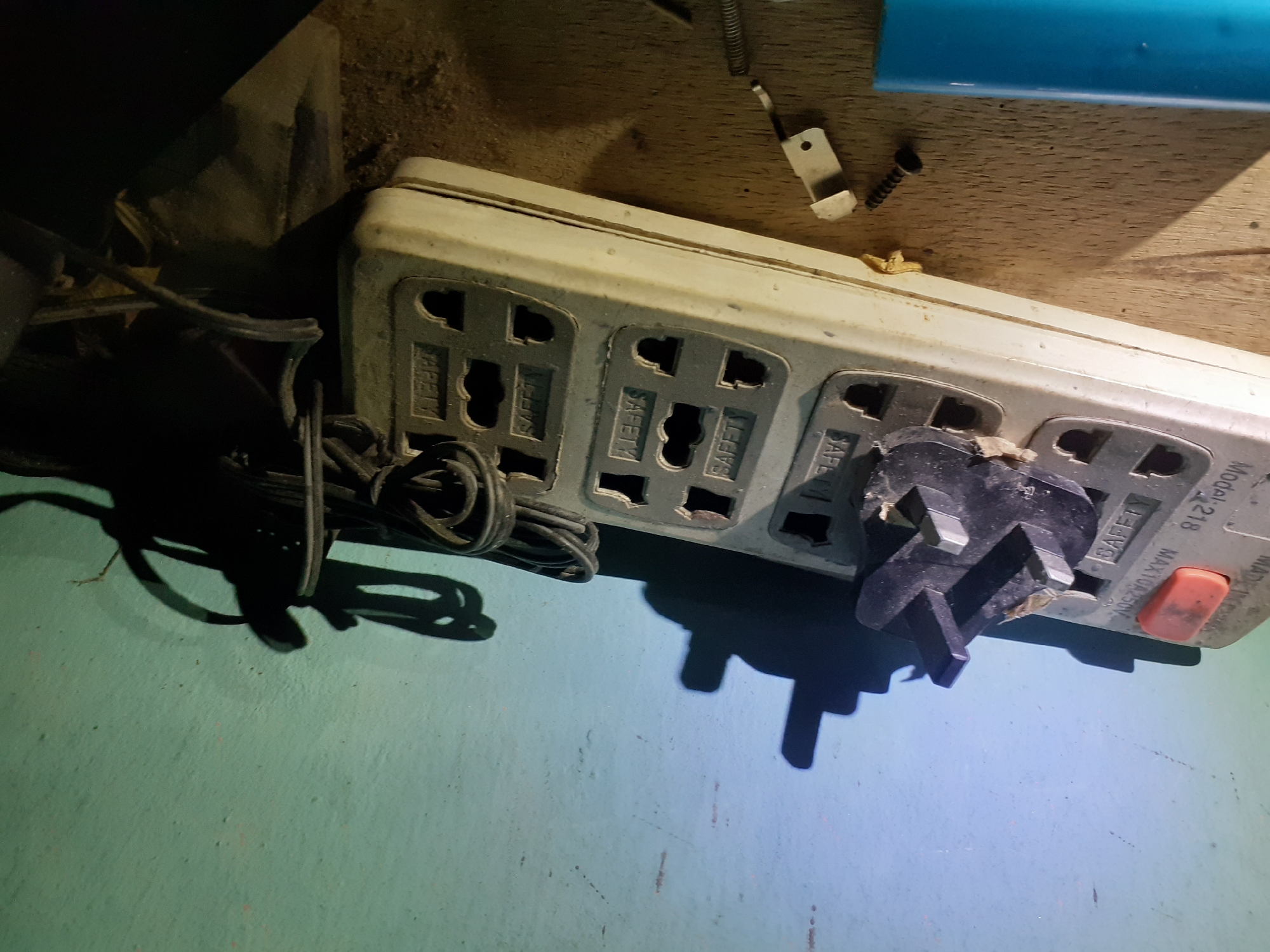 In an indoor environment, a close-up shot captures a white power strip with multiple outlets. A black, three-pronged plug is inserted into one of the outlets. The power strip is positioned on a wooden surface, likely a table or shelf. To the right, a coiled black electrical cord lies on the surface. In the background, a teal-colored wall is visible, with shadows cast upon it, suggesting a light source positioned to the left. A blue cylindrical object is partially visible in the lower left corner of the image. Scattered on the wooden surface are a black screw and a small metal bracket. The scene appears to be a static composition of electrical devices and related hardware.