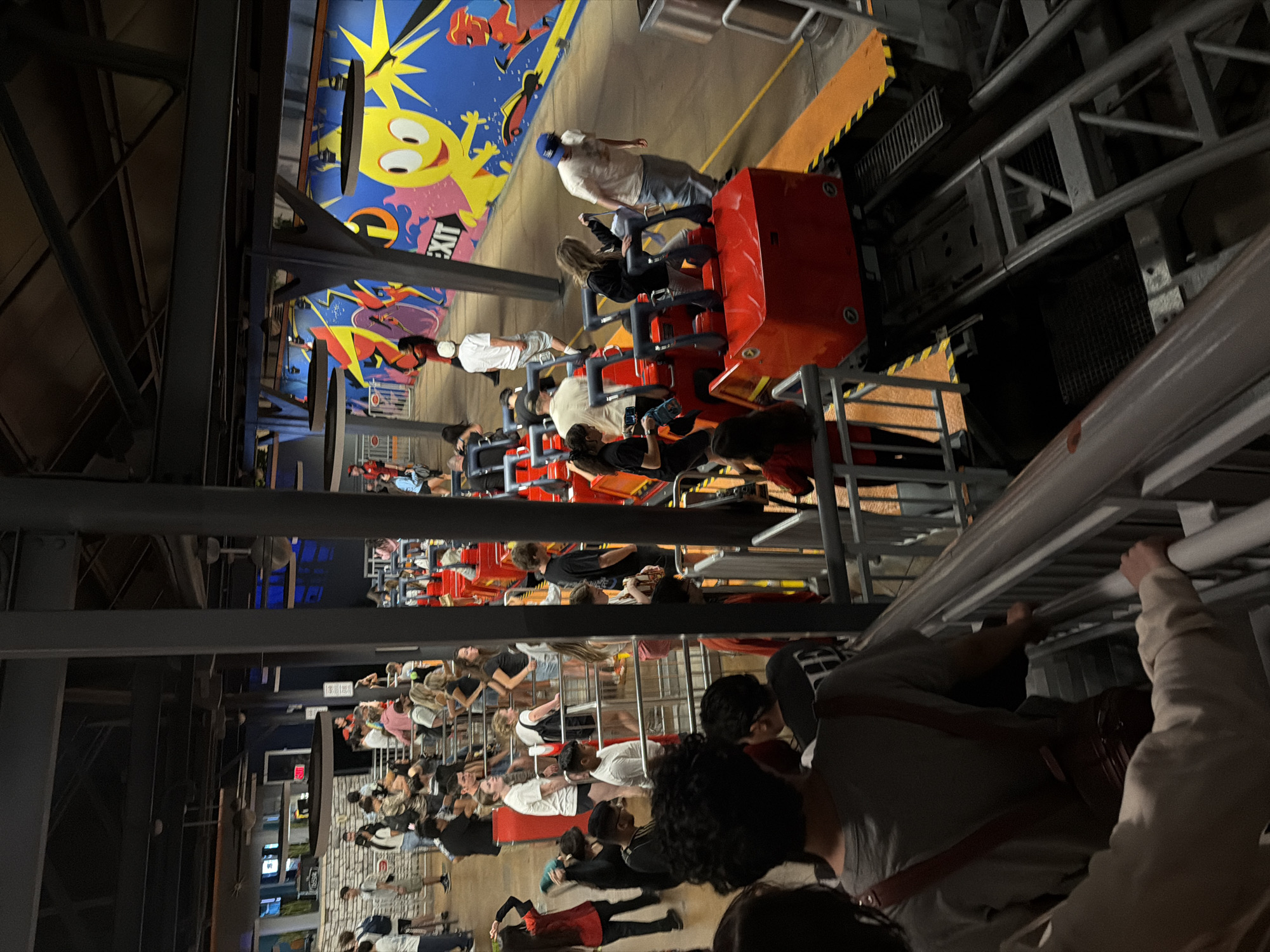 The photograph captures a queue of people waiting for a ride, likely a rollercoaster, at what appears to be a theme park attraction. A partially visible ride vehicle, red with rows of seats, is moving along the track. The surroundings are indicative of an indoor amusement area, with painted walls featuring cartoonish characters and steel support structures. The location is possibly Anaheim, United States of America, based on the theme park-like setting.