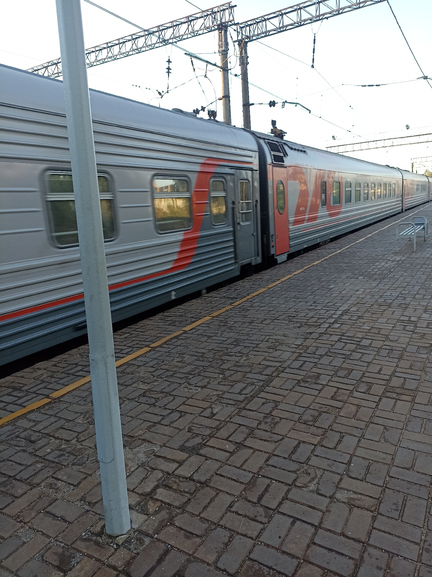The image captures a train at a station in Chekhov, Russia. The train is silver with red markings and appears to be moving or has just stopped, as indicated by the slight blur in the windows. The platform is paved with dark brickwork and a yellow line runs along the edge. A light-colored pole stands on the platform in the foreground. Overhead, power lines for the train are visible, supported by metal structures. The sky is clear and bright. There are no people visible in the image. A white bench is partially visible on the right side of the platform.