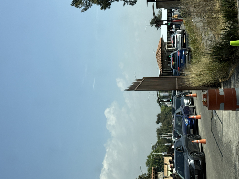 A street with a line of cars is visible in the lower right. Orange traffic cones are placed between the vehicles. A building with a tiled roof can be seen further down the road. To the left of the cars, a fence-like structure is visible, with streetlights lining the street. The sky is predominantly blue with some clouds visible. A tree is partially visible in the upper left corner.
