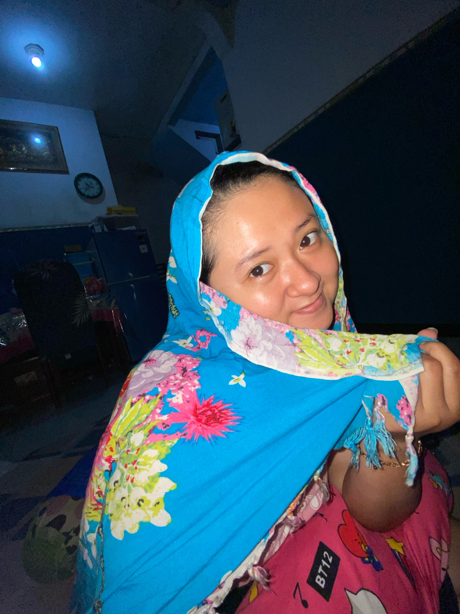 A medium shot portrait shows a woman from Indonesia in a dimly lit room, appearing to be at home in Gelam. She is looking at the camera with a slight smile and a playful glance. She is wearing a blue headscarf with a vibrant floral pattern of pink, white, and green flowers. The scarf is draped around her head and shoulders, and she is holding a corner of it with her right hand. Underneath the headscarf, she appears to be wearing a pink shirt with cartoon characters and the text "BT12". She has fair skin and dark hair.
The background is dark, with hints of a room interior. To the left, a framed picture hangs on a light-colored wall, and a round clock is visible below it. A light fixture with a blue glow is mounted on the ceiling above. To the right, a dark, angled surface suggests the presence of furniture or a wall structure. The overall lighting suggests it is nighttime, with the primary illumination coming from the ceiling light and the screen of the camera. The floor is covered with a patterned mat. The scene is intimate and casual, capturing a moment of personal expression.