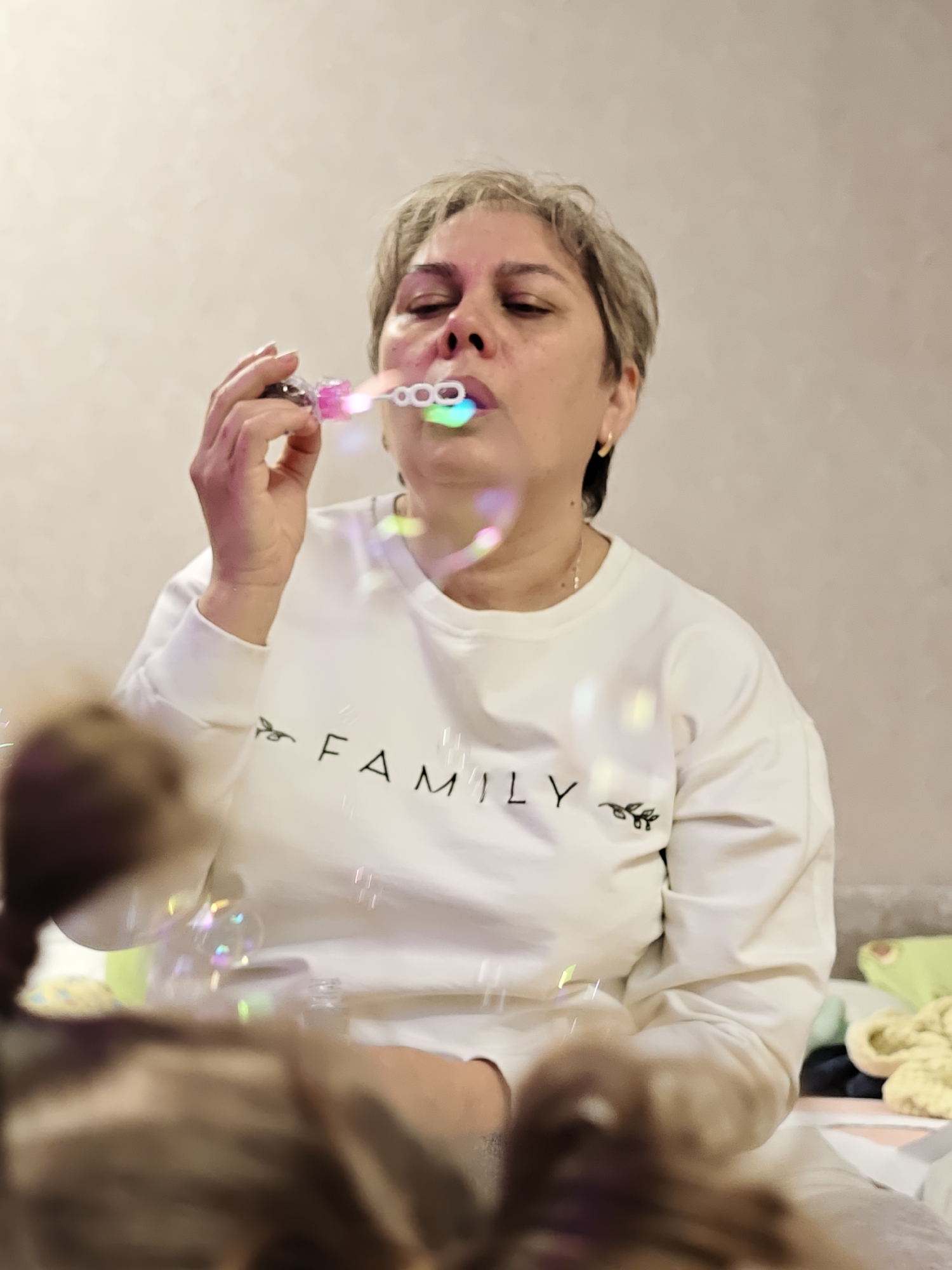 A woman with short, light-colored hair is blowing bubbles indoors. She is wearing a white sweatshirt with the word "FAMILY" printed on the front. She is holding a pink and white bubble wand to her lips and blowing, creating a stream of iridescent bubbles. The background is a plain wall, and the location is Chaikovsky, Russia.
