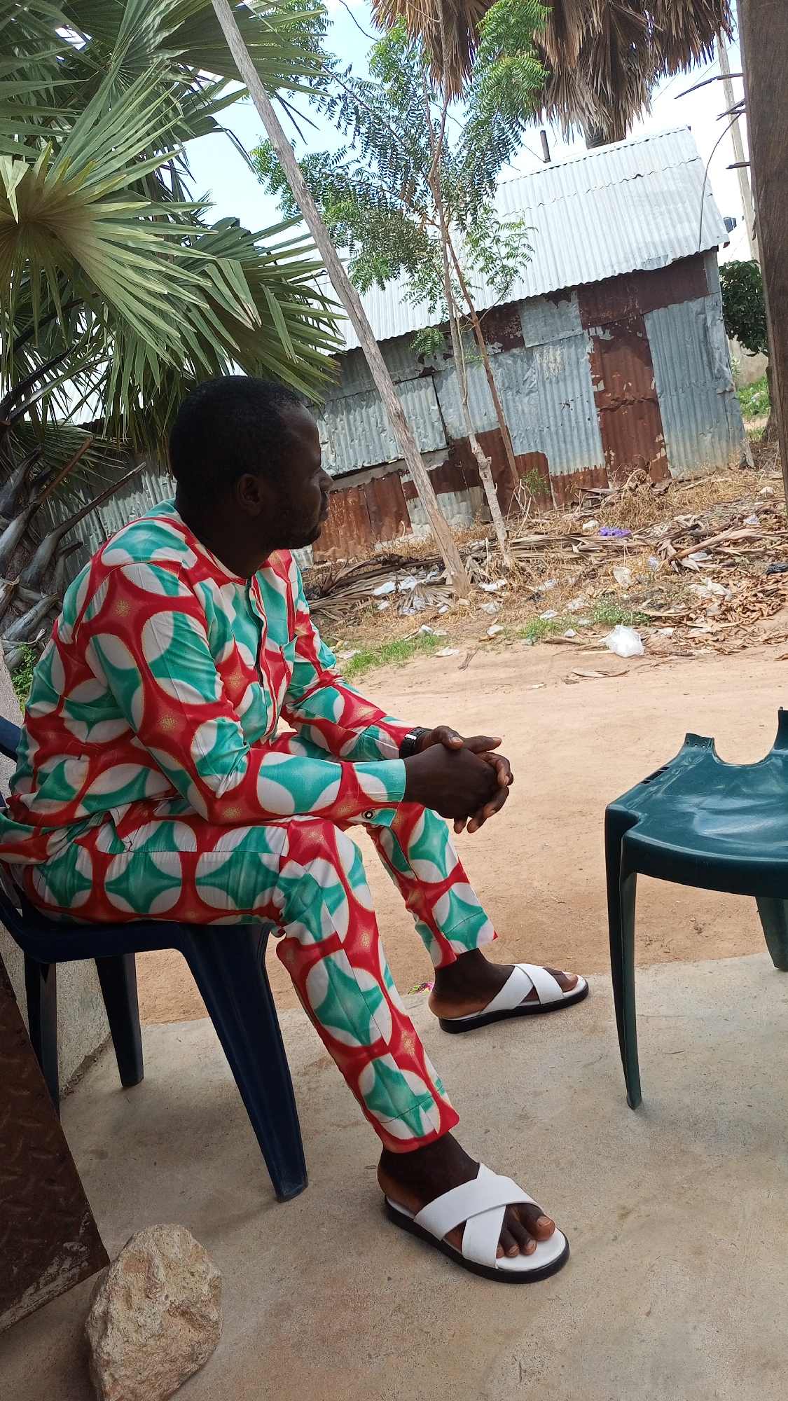 A man is sitting on a dark blue plastic chair, facing to his right. He is wearing a red, white, and teal patterned outfit, consisting of a long-sleeved shirt and pants. His hands are clasped in front of him, and he is looking off into the distance. He is wearing white slide sandals. The setting appears to be outdoors, with a sandy or dirt ground. In the background, there is a small building made of corrugated metal. There are also palm trees and other vegetation. The time of day appears to be daytime, with the sun casting shadows, suggesting it is likely midday or early afternoon. The weather seems warm and possibly dry, given the appearance of the ground and vegetation. There is no visible text in the image.