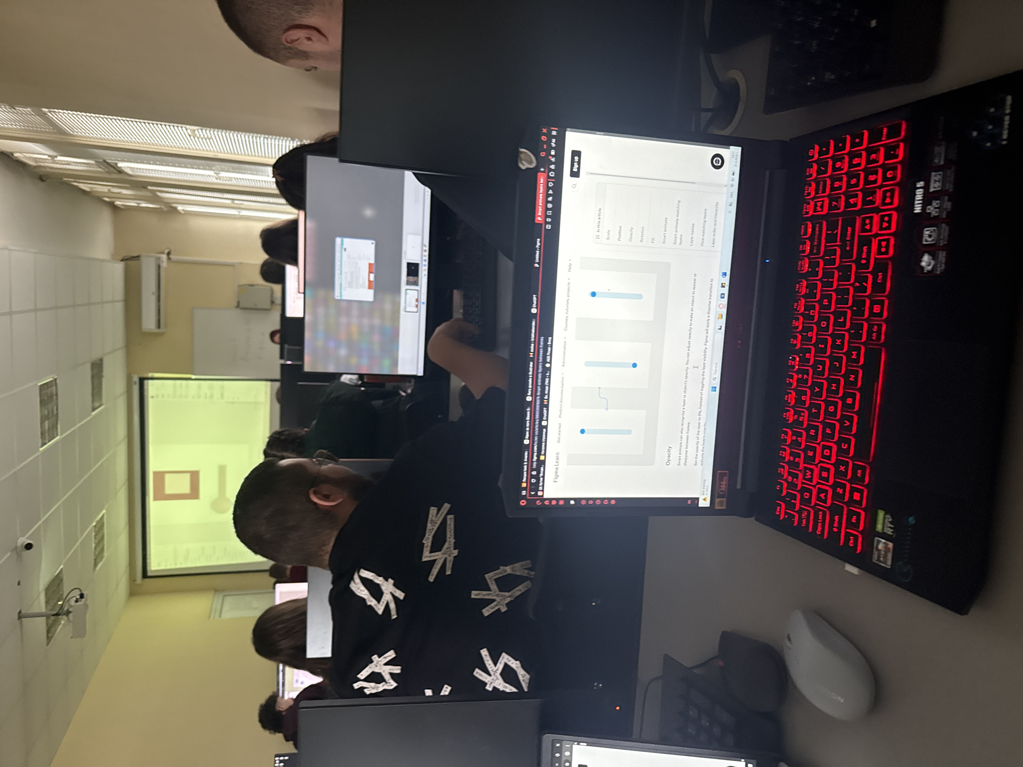 This rotated image depicts a busy computer lab or classroom in Plovdiv, Bulgaria, with multiple individuals engaged in computer work. In the foreground, a person wearing a black t-shirt with white abstract symbols is seated at a desk, intently looking at a desktop monitor. To their right, an Acer Nitro 5 laptop with prominent red backlit keys and a white ergonomic mouse is positioned. The laptop screen displays a web page, identified by the text "Figma Learn," which details design software functionalities like "Opacity," "Scale," "Position," and "Rotation," complete with adjustable sliders and textual descriptions about "Smart animate" and layer properties.
Behind this setup, other students are visible, also working on their computers. In the background, a large projection screen displays a simple graphic—a red square on a light background—suggesting a lesson or presentation is in progress. The room features fluorescent ceiling lights, a wall-mounted air conditioning unit, and whiteboards. A security camera is also visible on a side wall. The scene captures an educational or workshop environment focused on digital design or computer-related tasks. Notable visible text includes "NITRO 5" on the laptop, "Figma Learn," "Opacity," "Smart animate," and various keyboard labels like "Caps Lock," "Shift," and "Ctrl."